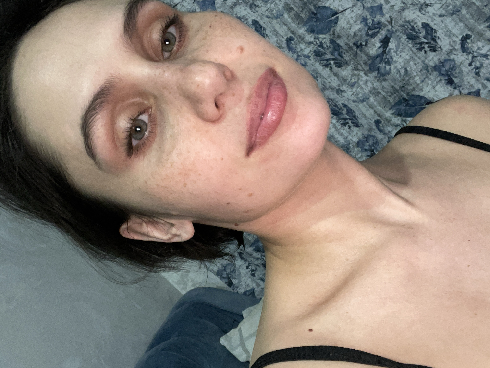 A woman with fair skin and freckles is lying down. Her eyes are hazel, and she has dark hair. She is wearing a black top with thin straps. The background is a blue and gray patterned fabric, possibly a blanket or sheet. There is no evidence to suggest this photo was taken in Izhevsk, Russia.