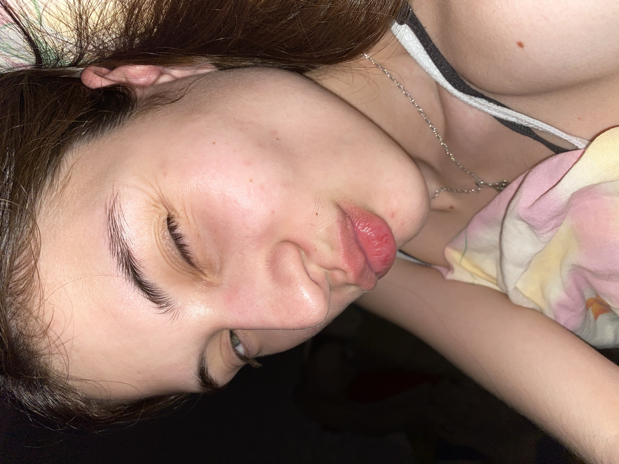 A close-up, angled shot features a woman's face, with her head tilted to the side and down. The top of her head is towards the top left of the frame, with her face angled down towards the bottom right. Her lips are slightly pursed. A silver necklace rests on her chest. Parts of a patterned fabric (possibly bedding or clothing) are visible, along with glimpses of skin and the edge of what appears to be a bra strap. The background is dark and out of focus. There is no information available to determine the location of the photo or that it was taken in Votkinsk, Russia.