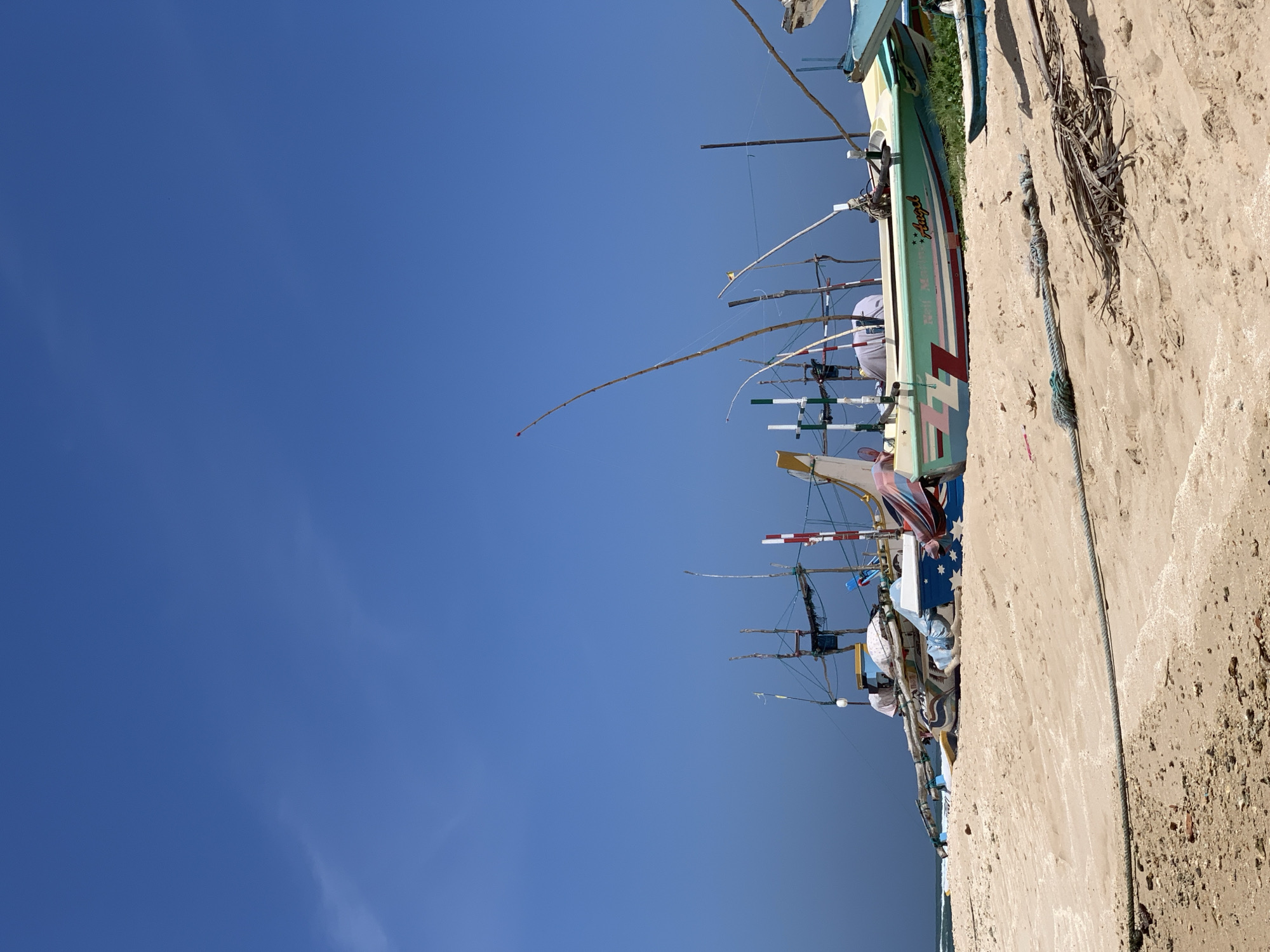 A scene in Mirissa, Sri Lanka, features several fishing boats resting on a sandy beach under a clear blue sky. The boats, varying in size and color, are arranged in a row, some partially covered by tarps. The foreground is dominated by the sand, with some dried palm fronds and a thick rope lying on the surface. The background shows the vast expanse of the sky and the distant ocean. The overall impression is one of a typical day at a coastal fishing village.