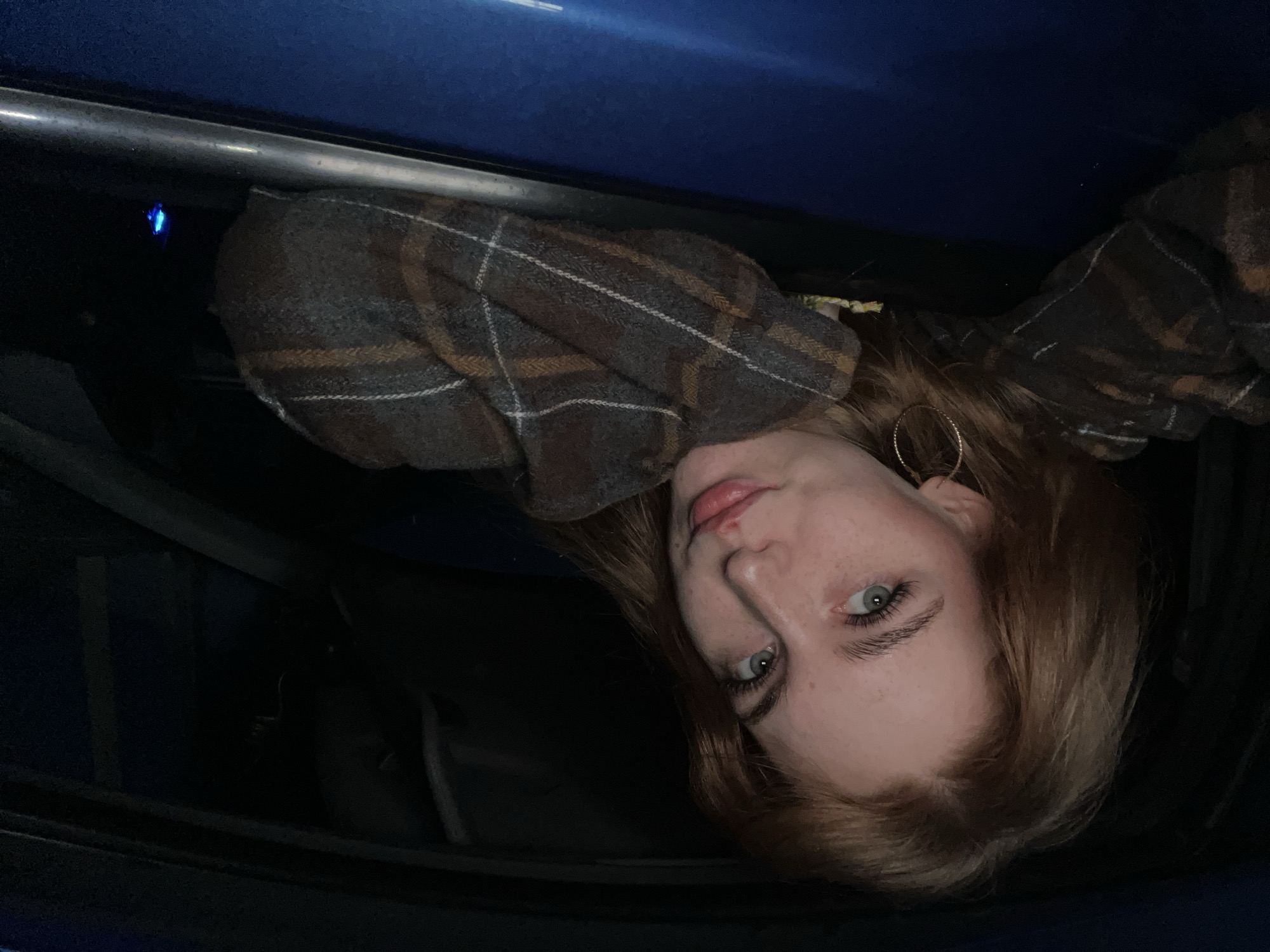 A young woman with auburn hair and light blue eyes is leaning out of a car window at night. She is wearing a plaid shirt and a hoop earring in her visible ear. The car is blue. The background is dark, with some faint blue light visible in the distance on the right side of the image, possibly from a streetlamp or another vehicle. The woman's pose suggests she is relaxed and looking towards the camera. There are no other people or animals visible in the image. The scene appears to be in an urban or semi-urban environment.