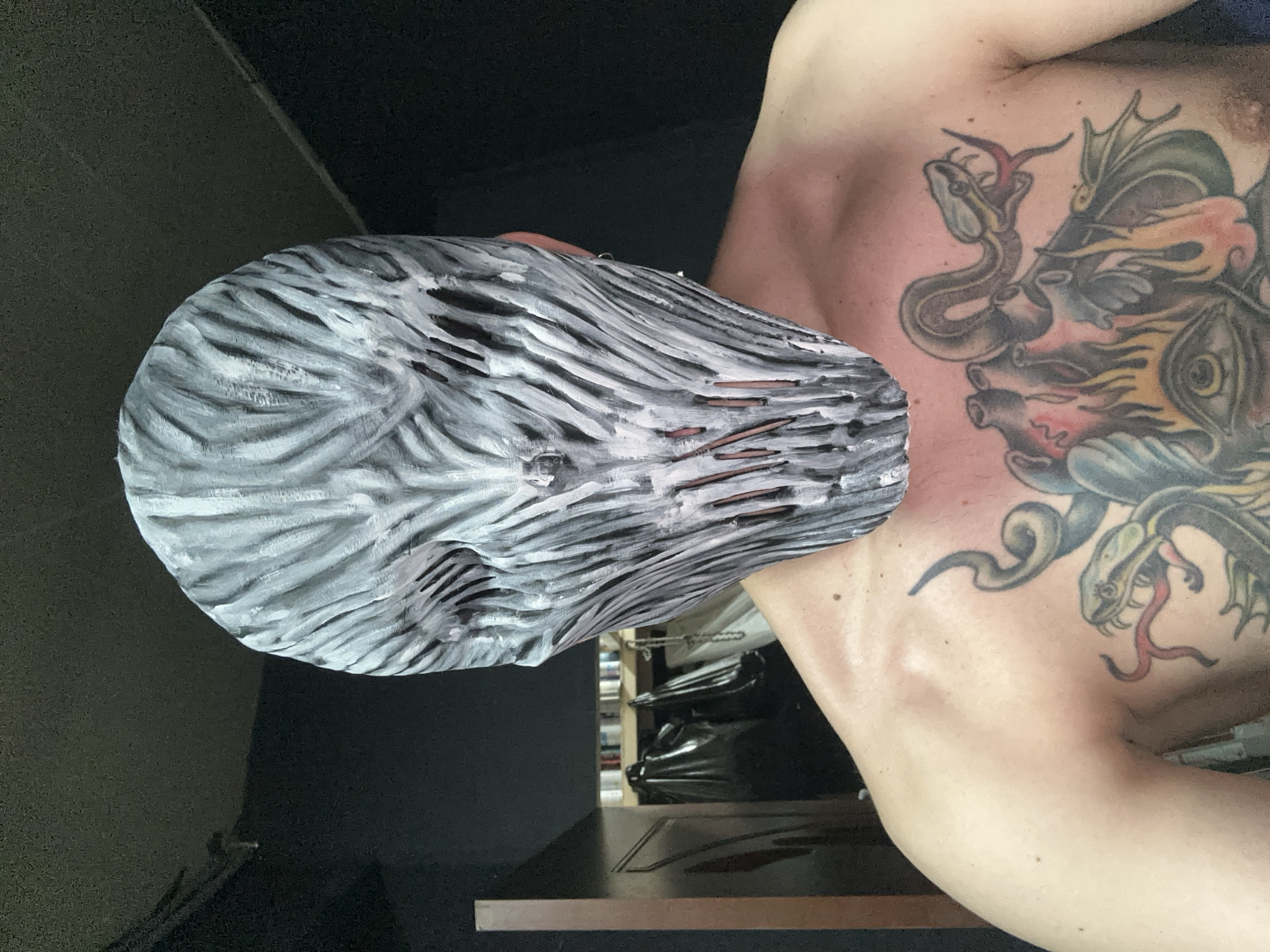 An individual, presumed male, is depicted in a frontal, waist-up view within what is stated to be a private indoor setting in Saratov, Russia. The individual wears a full-face mask, predominantly grey and white with a highly textured, vertically striated surface resembling organic fibers or muscle tissue. The mask features several narrow, dark vertical slits in the lower facial area. Below the mask, the individual's bare chest is extensively tattooed with intricate, multi-colored designs. The left pectoral area displays a green and brown snake with a red, forked tongue. The central and right pectoral areas feature a composite tattoo including a stylized anatomical heart, orange and yellow flames, a prominent eye, and dark, bat-like wings. The background is largely dark and out of focus. Visible elements on the left include a dark wooden furniture edge, a hanging black plastic bag, several stacked cylindrical objects (possibly cans) on a shelf, and a white chain. The individual is stationary, posing for the photograph.