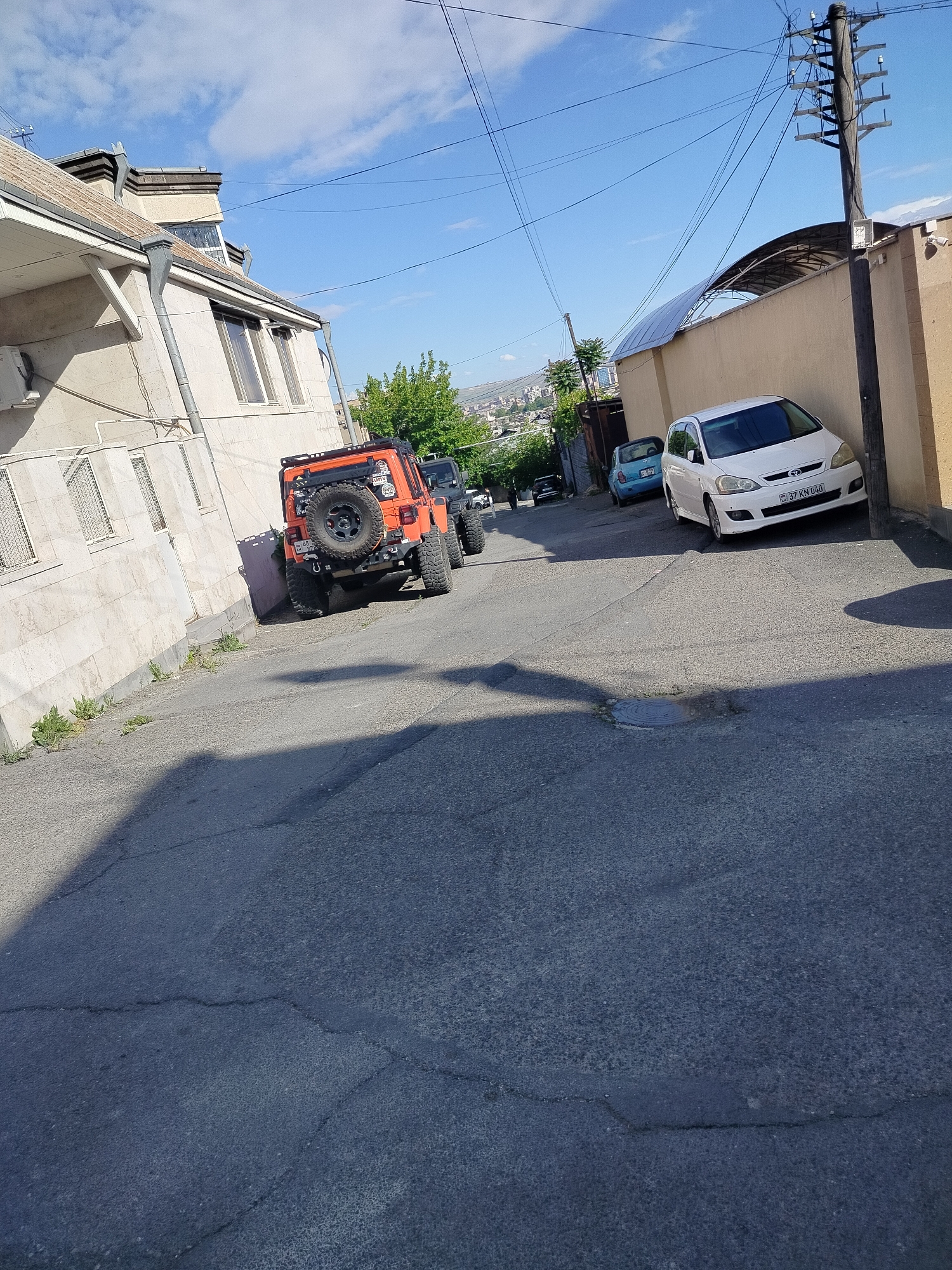 The image shows a narrow street in Yerevan, Armenia, with parked cars and buildings on either side. The street slopes upward into the distance. On the left is a light-colored building with multiple windows and a white-washed exterior. An orange Jeep with a spare tire mounted on the back is parked along this building. Further up the street is a black SUV. On the right side, a tan-colored wall extends along the street, next to a utility pole with power lines. A white car with the license plate "37 KN 040" is parked against the wall, with a light blue car behind it. The sky is blue with scattered clouds. The asphalt street has cracks and variations in color due to shadows and repairs.