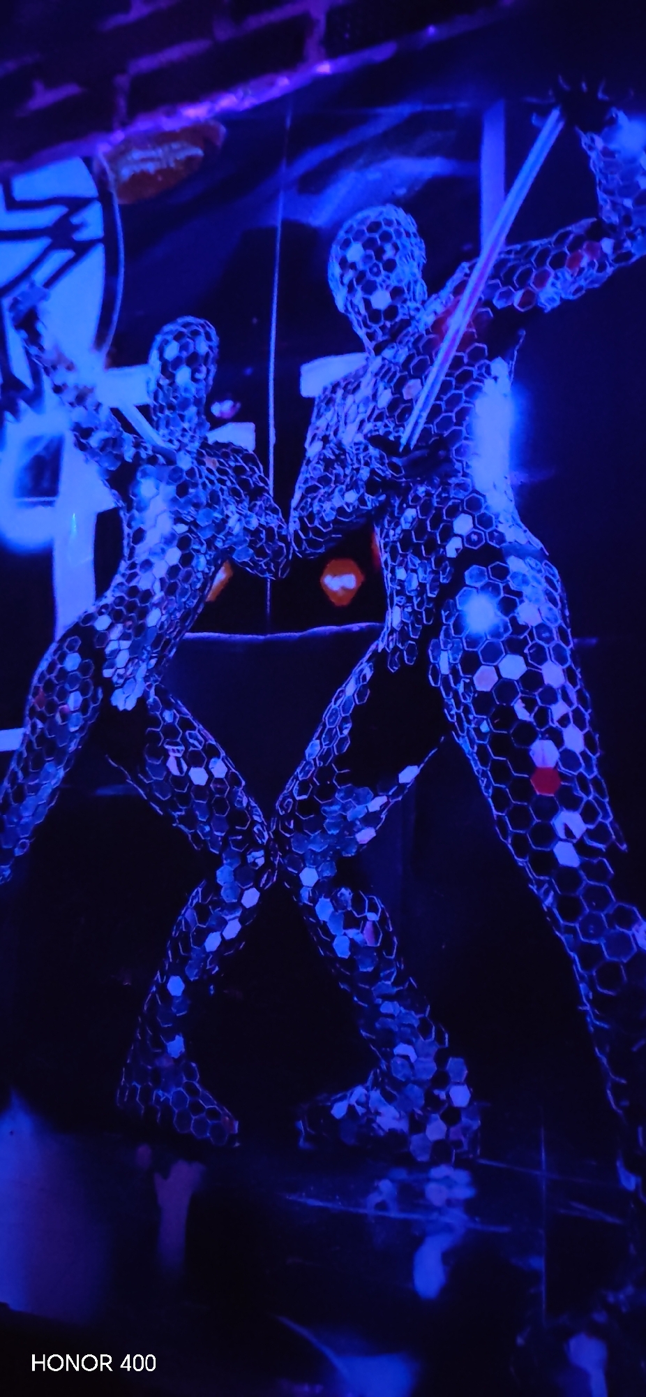 The image depicts two figures covered in a reflective, honeycomb-patterned material. They are illuminated by blue light in a dark environment. The figures appear to be mirrored, with one in the foreground and the other behind. The figures are holding objects resembling swords or batons. The scene is futuristic or theatrical in nature. The bottom left corner of the image contains the text "HONOR 400". There is no indication of Yakshur-Bod'ya, Russia, in the image.
