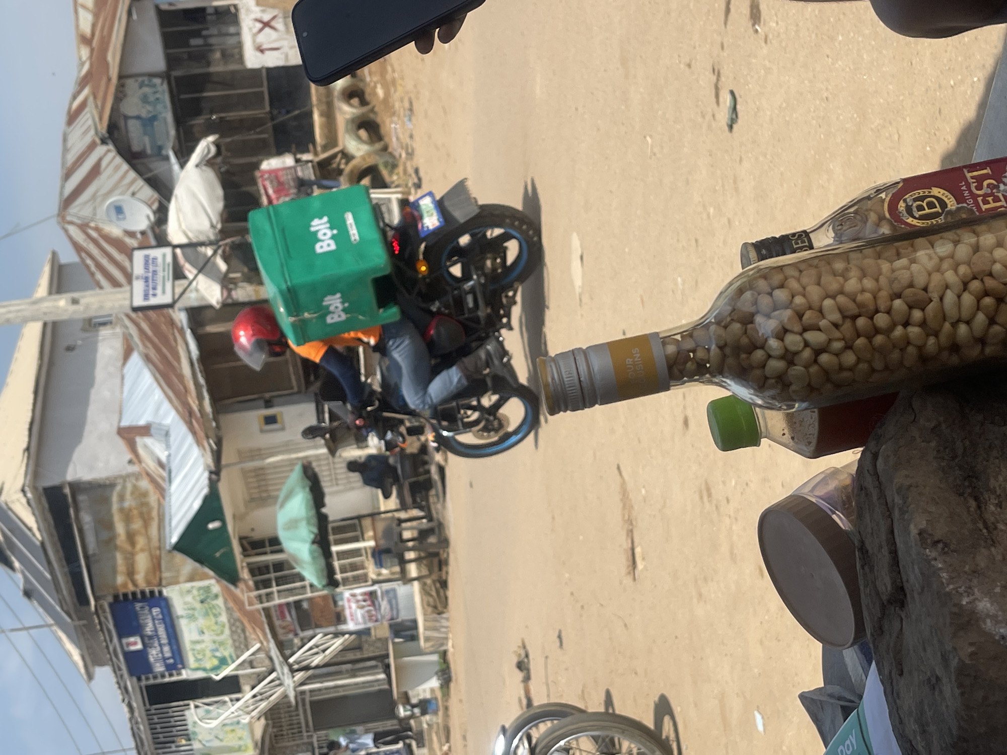 The image depicts a street scene in Abuja, Nigeria, with a focus on a Bolt delivery driver on a motorcycle. The driver, wearing a red helmet and an orange shirt, is operating a motorcycle equipped with a large green Bolt branded delivery box. The motorcycle has a blue rim on its rear wheel and a license plate. In the foreground, a variety of items are arranged on a surface, including a large glass bottle filled with what appear to be peanuts, a smaller bottle with a green cap, and a dark-colored container with a brown lid. In the background, buildings with corrugated metal roofs are visible, along with a blue sign that reads "WHITEHEART PHARMACY & MINI-MARKET LTD." Several people are present in the background, some standing and others seated, suggesting a commercial area. The ground is dusty, consistent with an outdoor urban environment.