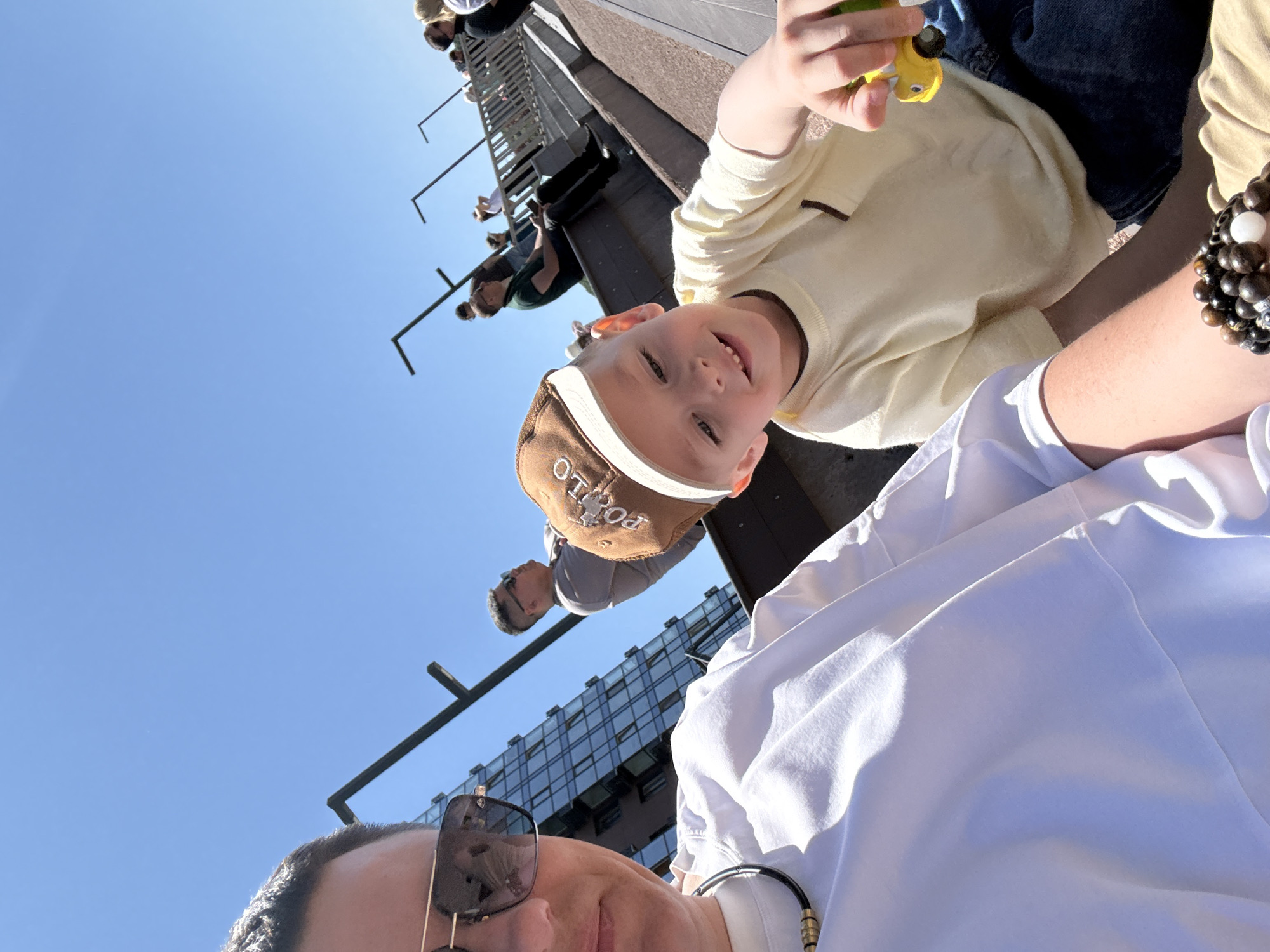 A low-angle photograph captures a man taking a selfie with a young boy. The man, wearing sunglasses and a white shirt, is partially visible at the bottom of the frame. The boy, wearing a brown "POLO" hat and a light yellow sweater, is closer to the camera and holds a yellow toy. Behind them, a building with a glass facade and other people on a structure resembling a ladder or tower are visible. The sky is blue and clear. The location is Vladivostok, Russia.