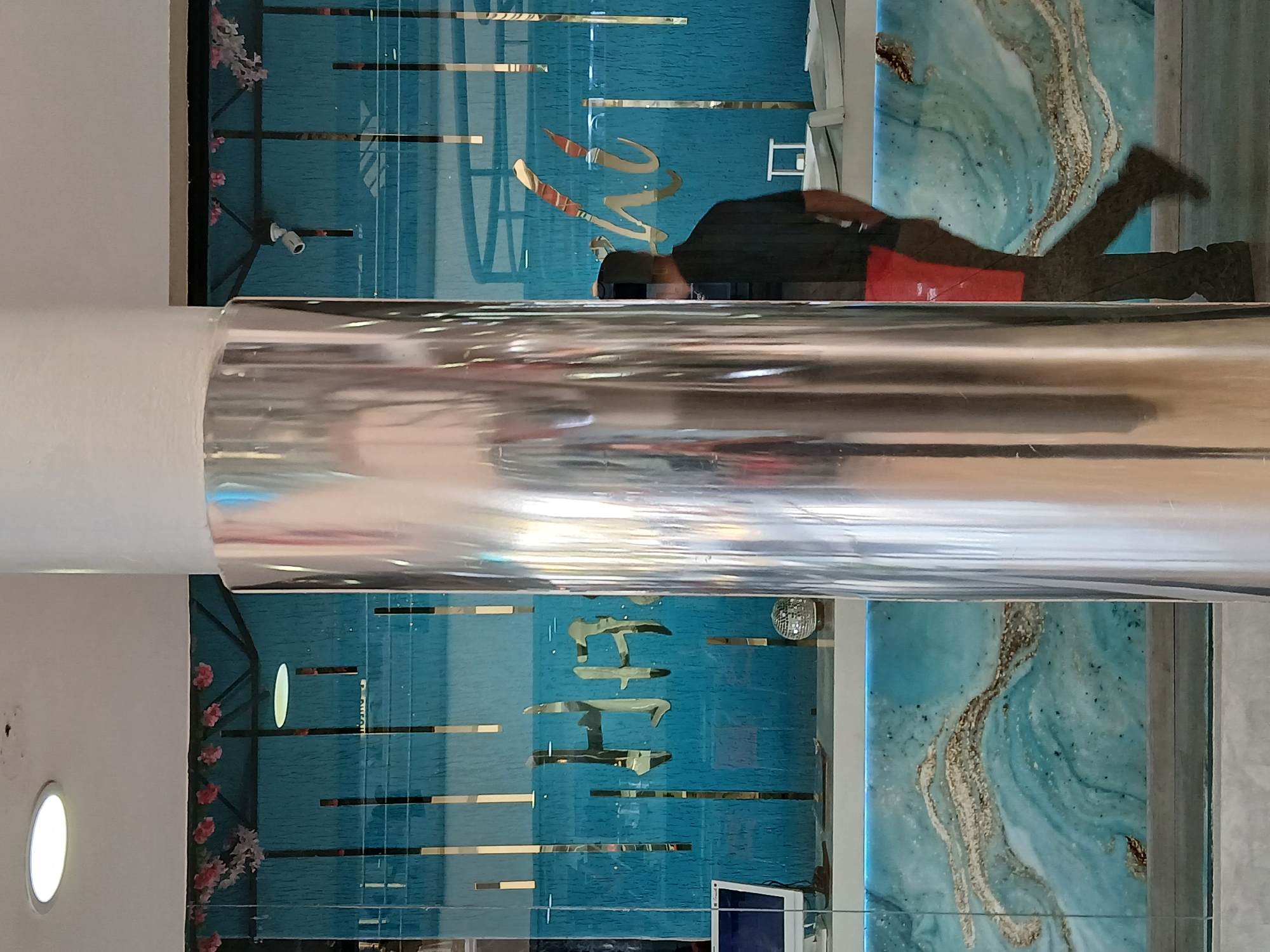 The image appears to be taken in an interior space in Ciudad Guayana, Venezuela. The main feature is a polished, cylindrical column reflecting the surroundings. Behind the column, there's a decorative wall. The upper section has an abstract ocean-themed painting in blue and white tones, along with golden lettering and horizontal lines. In front of the wall, there is a person wearing dark clothing with a red element, possibly walking past. The lower section of the wall has a similar blue and gold design with lettering, along with additional elements such as flowers, a security camera, a disco ball, and a television screen. The wall may be made of glass or another reflective material, amplifying the reflection of the environment. The scene is indoors.