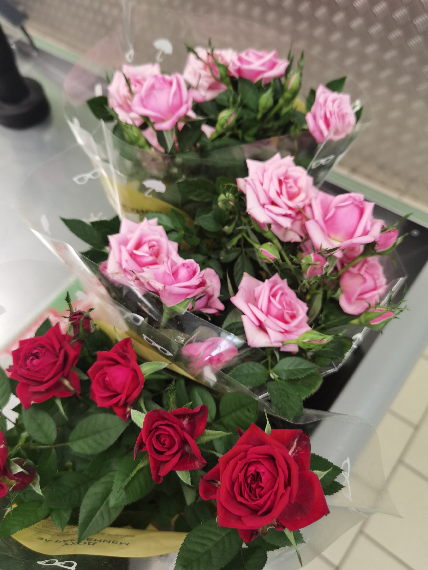The image shows two bouquets of roses, one with pink roses and the other with red roses. Both bouquets are wrapped in clear plastic packaging, and are placed on a metallic surface. The pink roses are in the upper part of the image, and the red roses are in the lower part. The scene appears to be indoors, possibly in a store or market, given the lighting and the nature of the objects. The background is out of focus but appears to be a light-colored, textured surface. There is no indication of the location being in Chaikovsky, Russia, or any specific city.