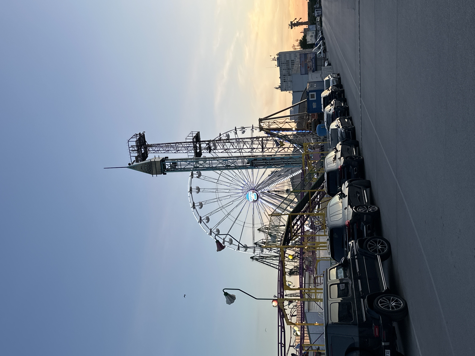 The image shows a Ferris wheel and a drop tower ride at an amusement park. Cars are parked along the side of the street, a row of mainly black SUVs, and a few more vehicles in the distance. The location is the amusement park in Varna, Bulgaria. The sky is blue and fades into orange/yellow near the horizon, suggesting a sunset. A bird is visible flying near the Ferris wheel. Buildings are visible in the distance. The lighting suggests it is late afternoon or early evening.