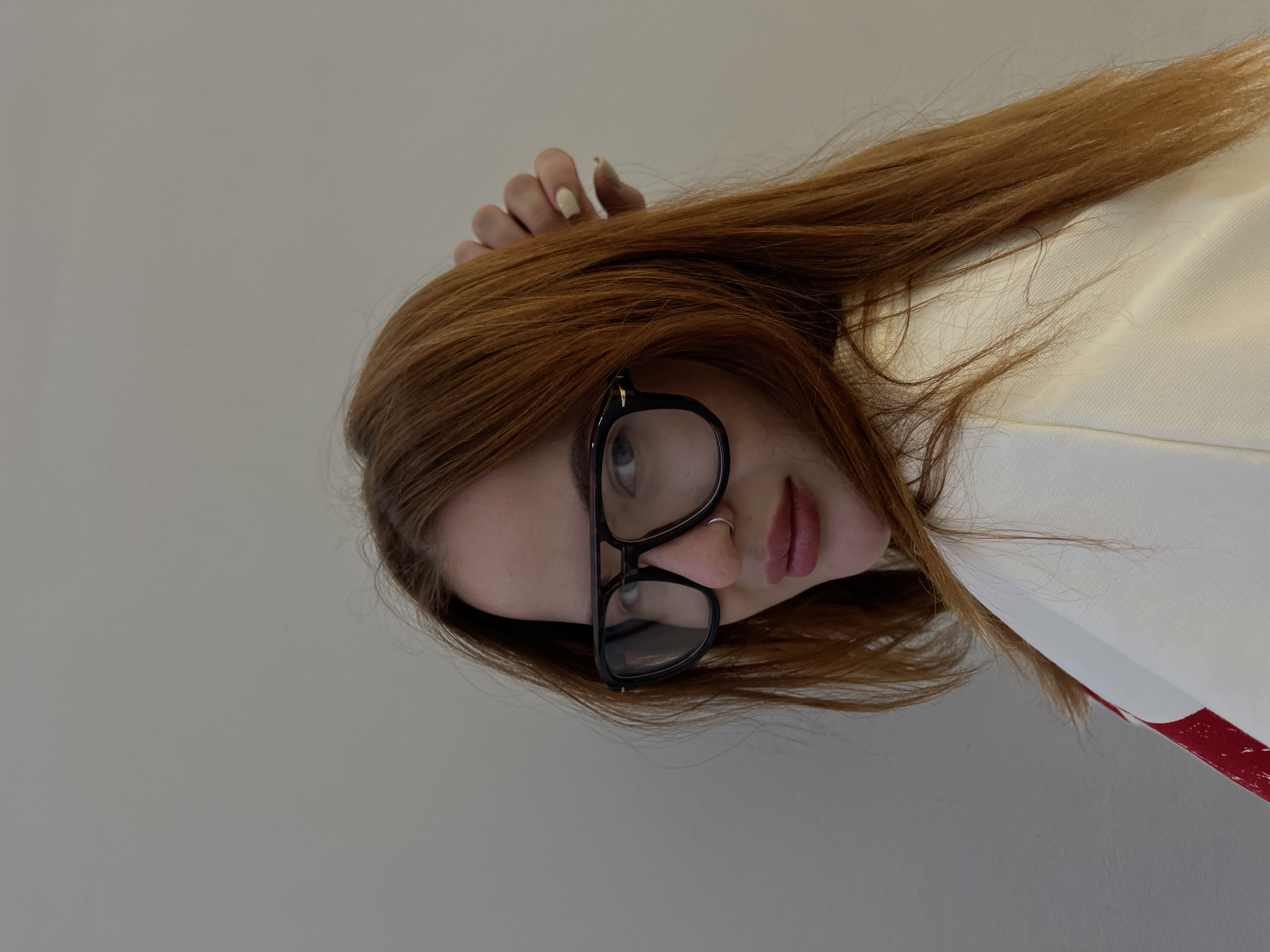 This is a rotated portrait photo of a young woman with long, reddish-brown hair. The image is oriented ninety degrees counter-clockwise, showing her head and upper torso from a slightly low angle against a plain, light grey background.
She is wearing large, black-framed glasses that obscure parts of her eyes, but her gaze appears to be directed upwards and to the right (relative to her own head). A small, silver hoop nose ring is visible on her left nostril. Her lips are a muted reddish-pink, slightly parted. One of her hands, with fingernails painted in a light, possibly cream or pale yellow color, is resting near her hair on the upper right side of the frame.
She is dressed in a white garment, which could be a shirt or jacket, featuring a prominent red stripe or design running vertically along its right side. The background is a simple, undecorated wall, suggesting an indoor setting. There are no specific cues regarding the time of day or weather, and her expression is neutral to slightly contemplative. No text is visible in the image.