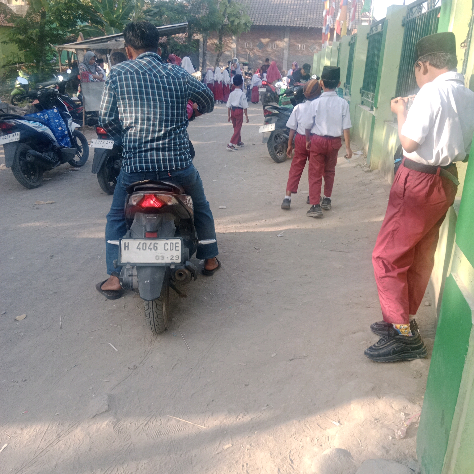 In Tegowanu Kulon, Indonesia, an outdoor scene unfolds with a person on a dark motorcycle in the foreground, facing away from the camera and towards a group of children. The rider wears a blue and white plaid long-sleeved shirt, blue jeans, and dark flip-flops. The motorcycle, also dark, has a license plate reading "H 4046 CDE" and a visible sticker with "03-29". Behind the motorcycle, several children in school uniforms, consisting of white shirts and red pants, are walking or standing. One child in the center is walking away from the camera, while two others are on the right, one looking towards the camera. The environment appears to be a dusty road or path, with trees and a building with a tiled roof visible in the background. A green fence runs along the right side of the frame. Additional motorcycles are parked or in motion further back in the scene.