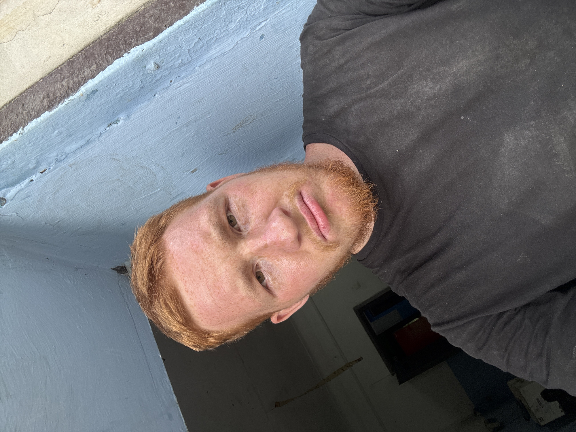 A close-up selfie shows a young man with ginger hair and a beard. He is wearing a black t-shirt. The background appears to be an outdoor urban environment, with blue and grey painted walls. The image quality is moderate. The man's expression is neutral.