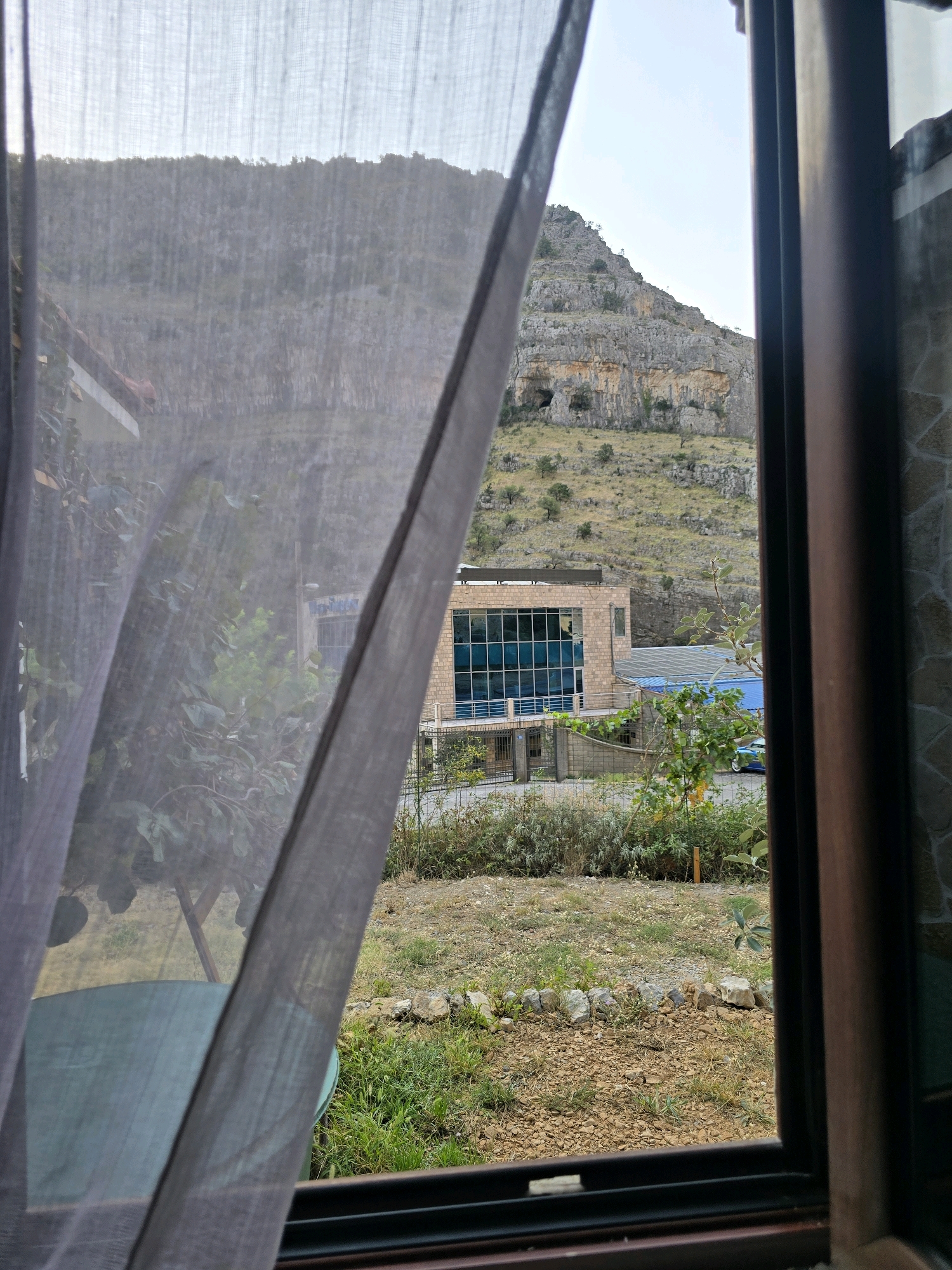 The image shows a view through a window, looking out onto a landscape in Podgorica, Montenegro. A sheer, grey curtain hangs on the left side of the frame, partially obscuring the view. In the middle ground, a modern building with a brick facade and large blue windows stands. In front of this building, there is greenery, including bushes and trees, and a small fence. To the right, a section of the window frame, made of dark wood, is visible, along with some stonework on the outer wall of the building from which the photo is taken. The background is dominated by a rocky, sparsely vegetated mountainside, with some trees and shrubs scattered across its slopes. The sky is a pale, overcast blue. The ground in front of the building appears to be a mix of gravel and grass. A light-colored car is parked near the building.
