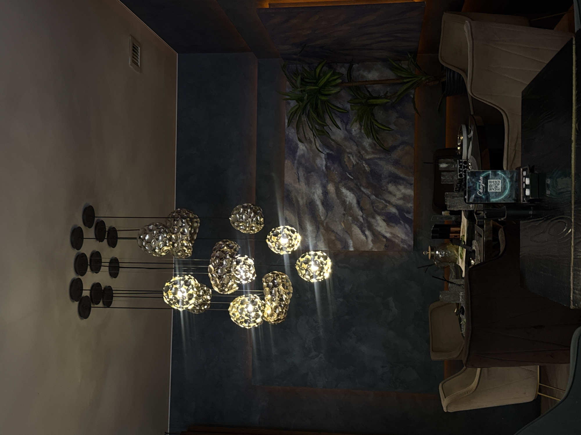 This is a medium-shot, eye-level, indoor photograph taken in the city of Unknown, Unknown. The scene depicts a dimly lit restaurant dining area. Dominating the upper portion of the frame is a cluster of decorative pendant lights, each with a unique, textured, spherical shade that emits a warm, yellow glow. The lights are suspended from circular ceiling fixtures by thin wires.
In the middle ground, several tables are set with glassware, plates, and other dining accoutrements. One table has a decorative vase and what appears to be a small device displaying a QR code. Surrounding the tables are plush upholstered chairs in neutral tones. To the right, a tall, potted palm plant adds a touch of greenery to the dark, textured walls. The walls themselves feature a marbled or abstract patterned wallpaper in shades of grey and blue. The overall ambiance is elegant and modern. There are no people visible in the image.
