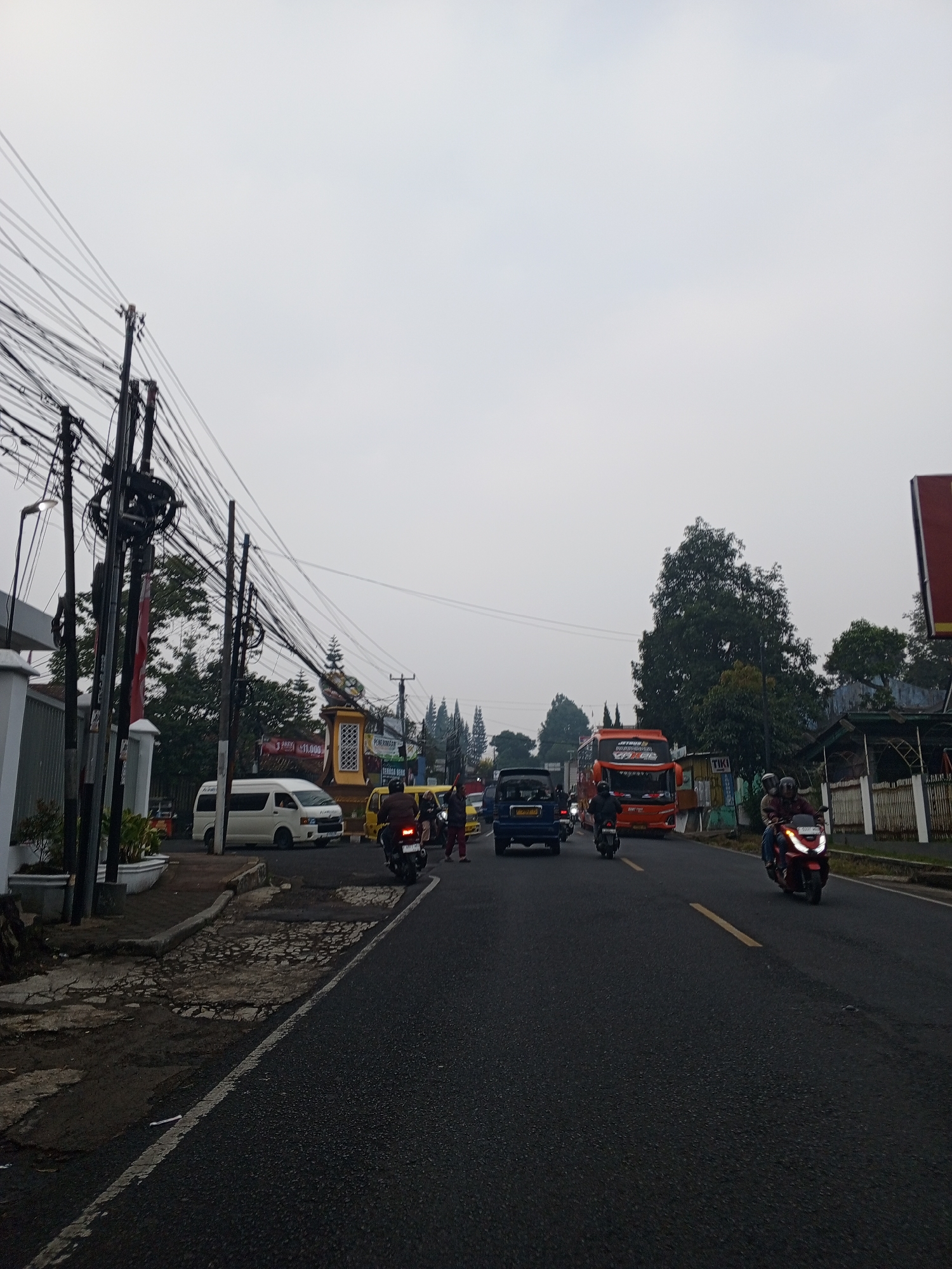 The image depicts a street scene with a moderate amount of traffic on an overcast day. In the foreground, the asphalt road is visible with a white line marking the lane division. Several vehicles are present, including a red scooter with two riders, a blue minivan, and a white van. A large orange bus is seen in the middle ground, along with other vehicles and motorbikes. There are numerous utility poles with a dense network of electrical wires on the left side of the road, characteristic of many urban and semi-urban areas. Trees line the right side of the road, and some buildings and structures are visible in the background. The overall impression is that of a busy road in a tropical or subtropical climate, possibly in Southeast Asia given the presence of the bus livery. The location is identified as the city of Dambulla, Sri Lanka.