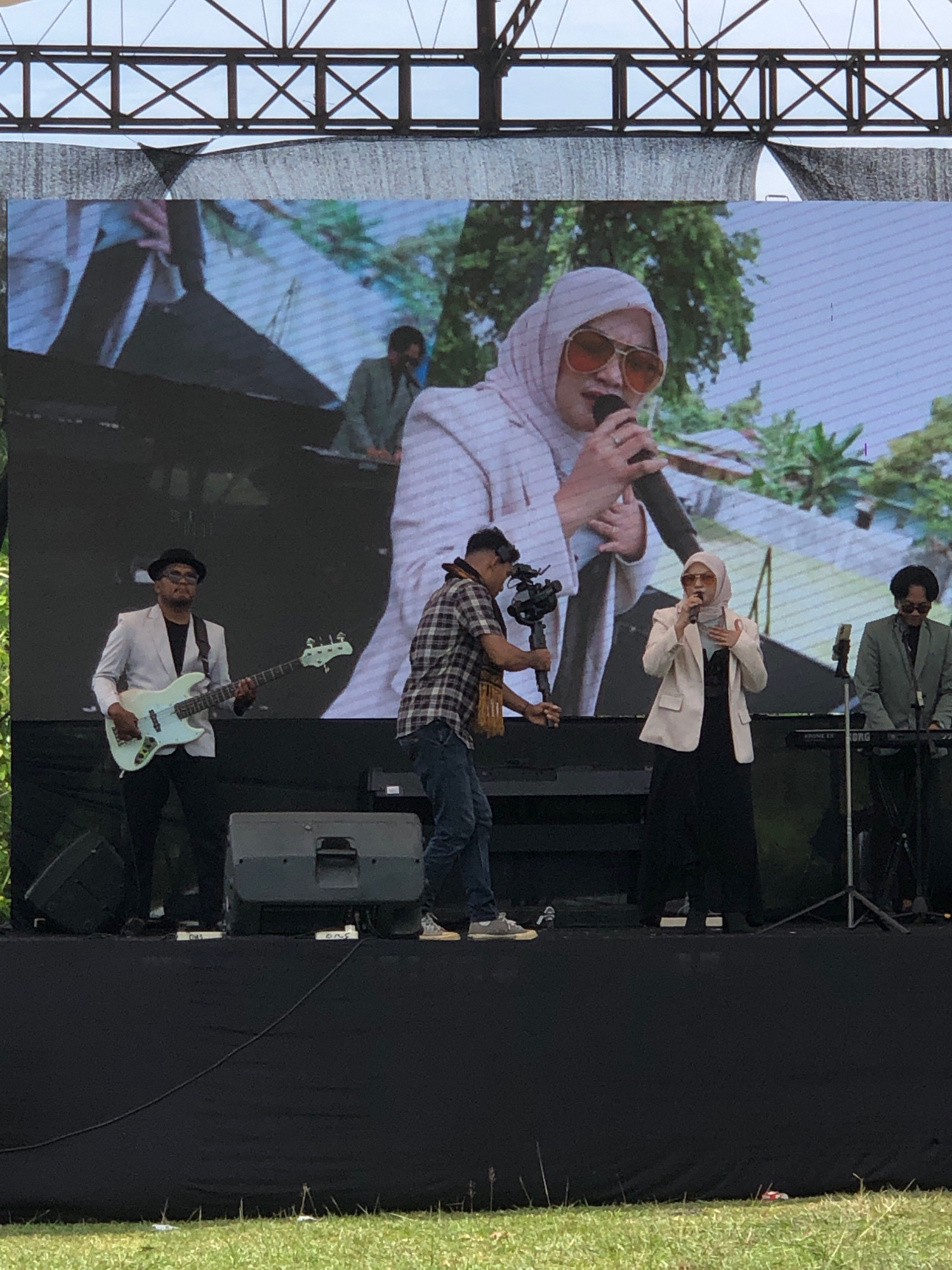 This is a photograph taken from a low angle, outdoors, likely at a music festival or concert in Lhokseumawe, Indonesia. The foreground is dominated by a black stage draped with fabric, with some speakers visible. Behind the stage, a large LED screen displays a performance, featuring a woman in a white hijab and sunglasses singing into a microphone. She is wearing a white blazer over a dark outfit. To her left, a man in a plaid shirt and jeans operates a camera gimbal. On the far left of the stage, a musician wearing a light-colored blazer and a hat plays a light blue bass guitar. To the right of the singer, another musician in a green suit plays a keyboard. A metal truss structure is visible above the screen, suggesting an outdoor venue. The overall atmosphere appears to be a live musical performance.