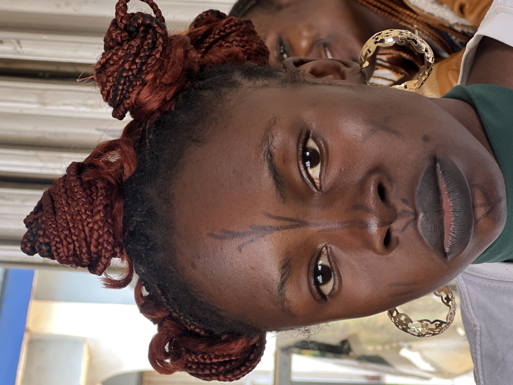 The image is a close-up portrait of a young woman with dark skin, styled hair, and prominent facial markings. She is wearing large, ornate gold hoop earrings. Her hair is braided in a deep red-brown color and styled into several top knots with some loose tendrils. On her forehead, there are dark, vertical markings resembling a stylized tree or a crest. Similar dark markings are also visible on her cheeks. She has dark lipstick applied to her lips. Her eyes are looking directly at the camera.
The background is somewhat blurred but appears to be an outdoor or semi-outdoor setting. The lighting suggests it is daytime. In the background, another person with braided hair is visible, smiling. The overall setting appears to be casual. The image is a portrait that emphasizes the woman's features and cultural adornments. The mention of Jos, Nigeria, in the prompt suggests the context of the image relates to the cultural landscape of that region.