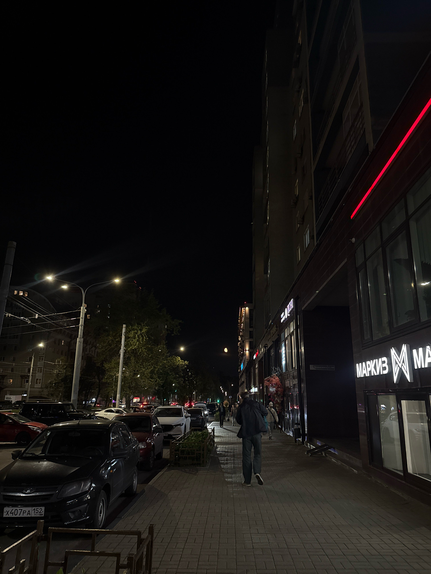 The image depicts a street scene at night in Nizhny Novgorod, Russia. A man walks away from the camera on a paved sidewalk, which is illuminated by streetlights. To the right of the sidewalk is a modern building with large windows and illuminated signage. One sign reads "МАРКИЗ". To the left of the sidewalk, cars are parked along the street, and trees line the opposite side of the road. The sky is dark, indicating it is nighttime.