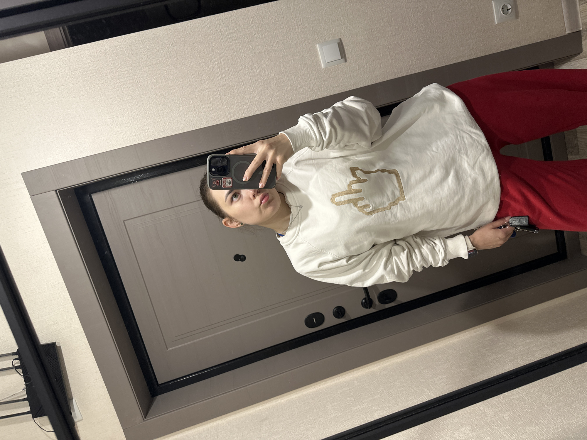 A young woman takes a mirror selfie inside an apartment in Perm, Russia. She stands, reflected in a large mirror, holding a dark smartphone in her right hand, positioned for the photo. Her left hand holds a set of keys with a dark fob. She wears a white long-sleeved sweatshirt featuring a prominent golden-colored graphic of a hand making an "up yours" gesture, along with red loose-fitting pants and a silver heart-shaped pendant necklace. Her dark hair is pulled back, and she has a subtle earring in her visible left ear. The background, reflected behind her, reveals a light gray-brown apartment door with black trim, equipped with a peephole, three black circular locking mechanisms, and a black handle. Above the door, a black Wi-Fi router with three antennas is mounted on the wall. The surrounding walls are light-colored with a subtle texture. A white rectangular light switch is visible on the wall to the right, and a white electrical outlet is positioned lower on the far right.