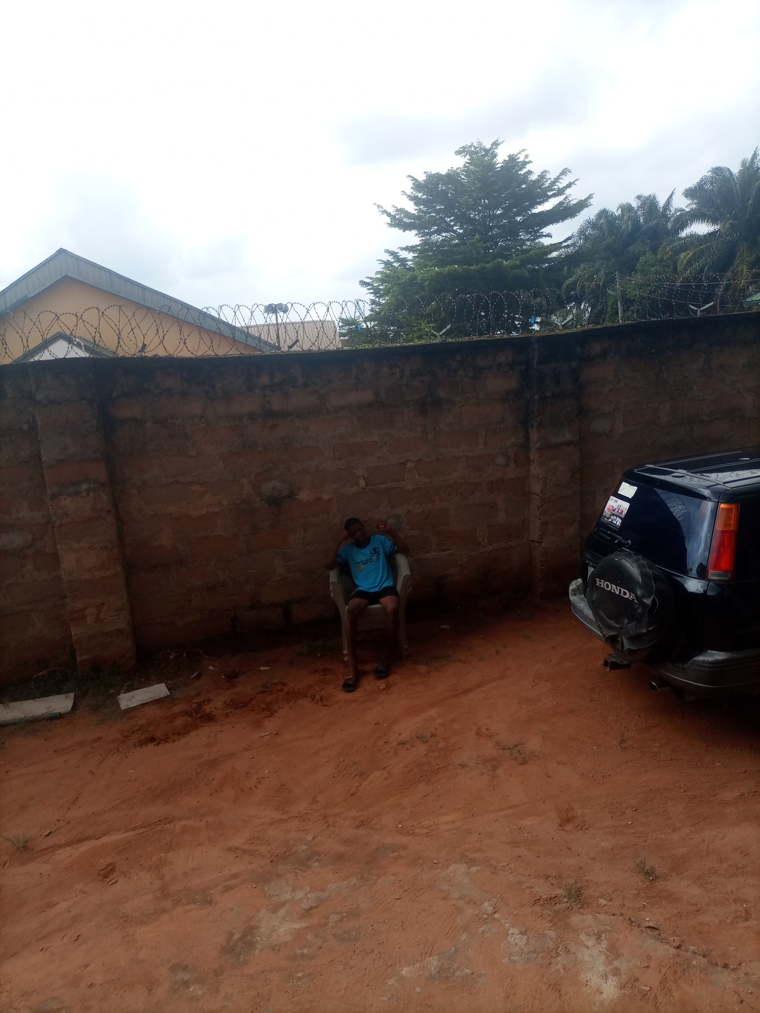 A young Black male sits on a plastic chair against a brick wall in Nkpor, Nigeria. He is wearing a blue t-shirt and black shorts, and is resting his head on his hand. A black SUV is parked to his right, with a spare tire cover that reads "HONDA". The ground is covered in red dirt, and there is a barbed wire fence visible above the wall. The sky is overcast.
