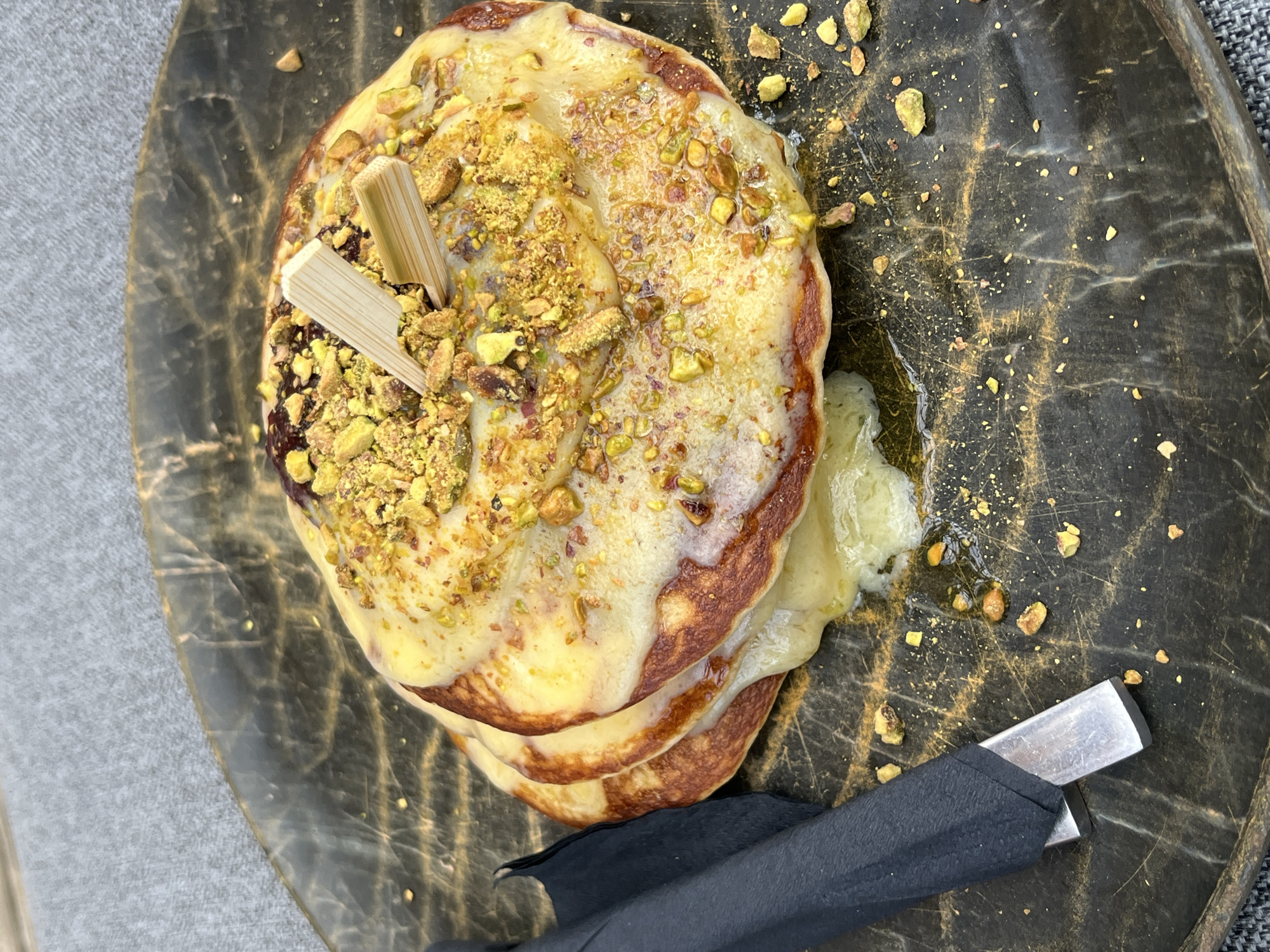 A close-up, overhead shot shows a stack of three pancakes topped with a creamy yellow sauce and chopped pistachios. Two small wooden skewers are inserted into the top pancake. The pancakes are on a dark, marbled plate. To the left of the plate, a black napkin is wrapped around a metallic utensil. The scene is set against a grey, textured background. The image is dated August 17th, 2023, and is geotagged to Thassos, Greece.