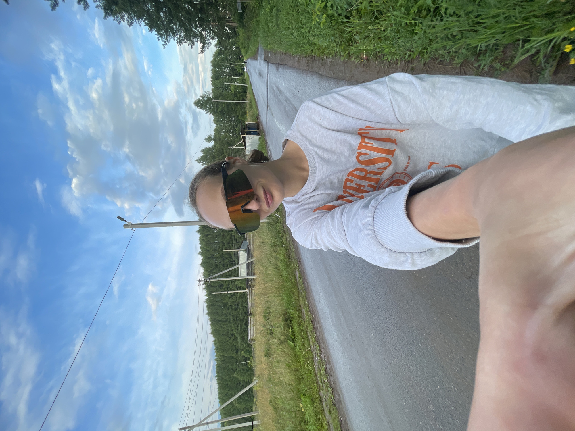 A young woman with blonde hair pulled back, wearing large, reflective sunglasses and a light grey sweatshirt with orange lettering, is taking a selfie on a wet, paved rural road. She is standing on the side of the road, which is bordered by tall green grass and trees. In the background, there are utility poles with wires, a dense line of trees, and a small, weathered building. The sky is cloudy with patches of blue. The scene appears to be set in or near the village of Podshivalovo, Russia.