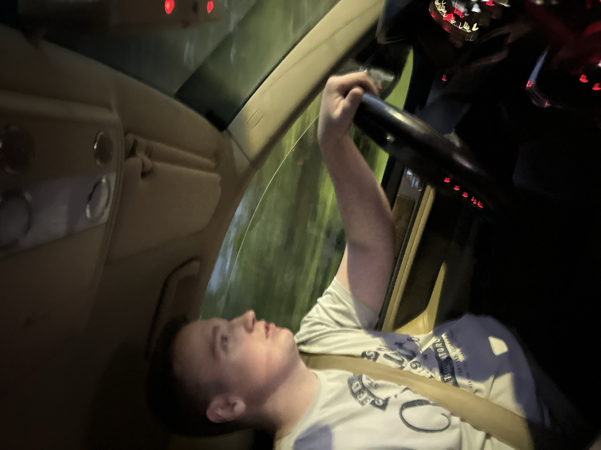 A passenger is reclined in the driver's seat of a car, holding the steering wheel. They are wearing a seatbelt and a white t-shirt with blue text. The car's interior is tan. Through the windshield, blurred lights and foliage suggest the car is moving at night. The location is unknown, and there is nothing in the image to indicate it is Nizhny Novgorod, Russia.