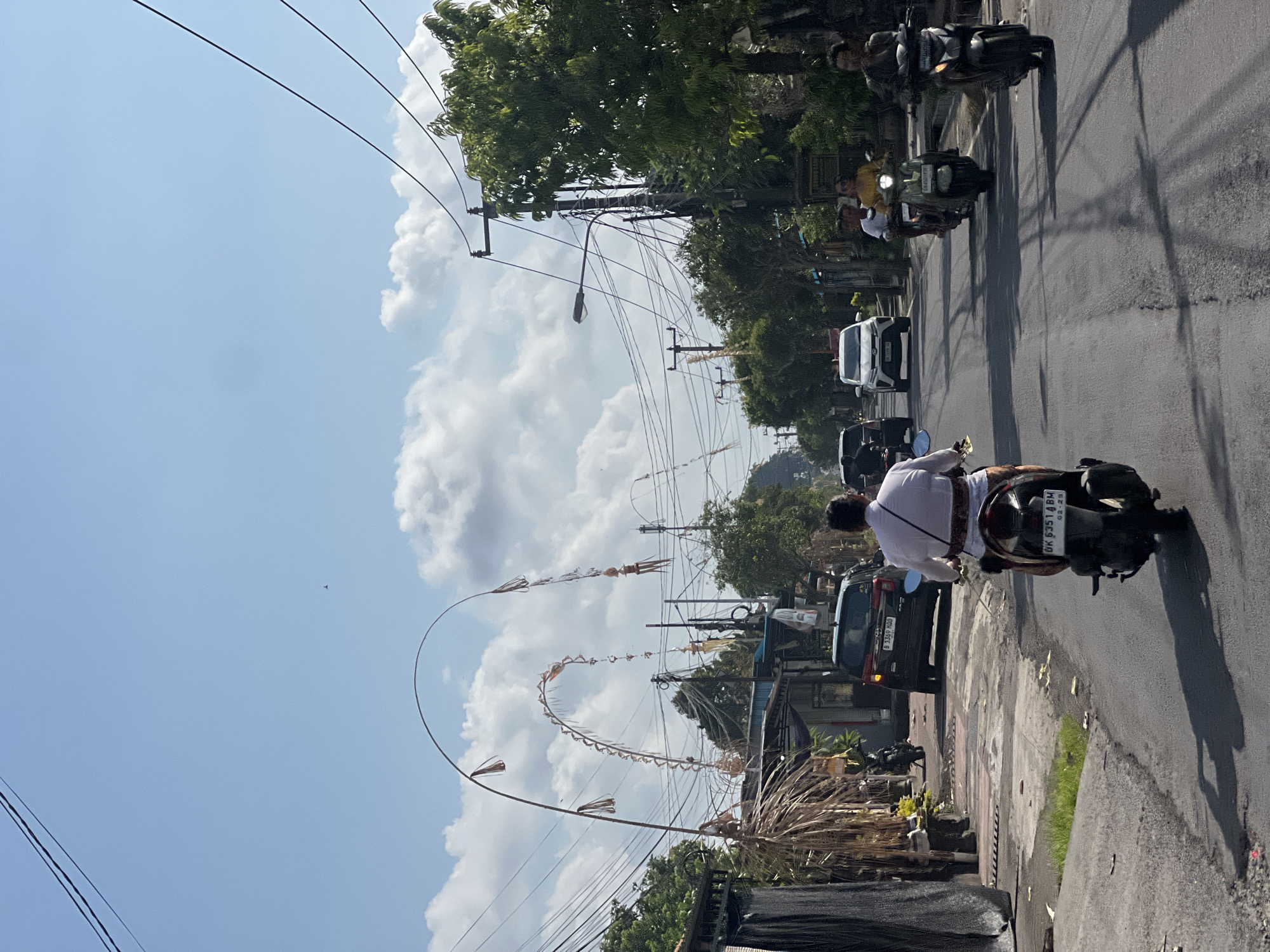 The image shows a street scene in Sukawati, Indonesia, on a bright, sunny day with scattered clouds. Several motorcycles and a car are visible on the asphalt road. A man in a white shirt and patterned skirt rides a motorcycle in the foreground, facing away from the camera. To his left, a dark-colored car is parked. On the right side of the road, two more motorcycles are riding towards the viewer, with passengers on the back. Buildings and lush green trees line the sides of the street. Decorative arched bamboo poles with hanging ornaments are prominent, suggesting a cultural or festive event. Utility poles and overhead wires are also visible, indicating the presence of infrastructure. The overall atmosphere is one of everyday life in a tropical urban environment.