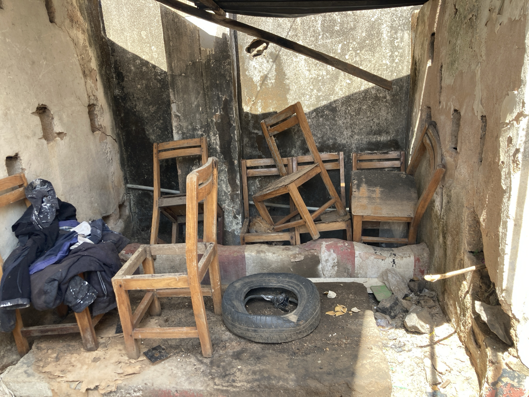 The image shows a dilapidated indoor space, possibly a storage area, in Jos, Nigeria. Walls are crumbling, revealing exposed brick and plaster. Several wooden chairs, some broken or disassembled, are scattered throughout the room. A tire lies on the floor in the center of the room. A pile of dark clothing is draped over one of the chairs. Debris, including broken pieces of wood and rubble, is visible on the floor. The room is dimly lit, suggesting a lack of natural light. There are no people visible.