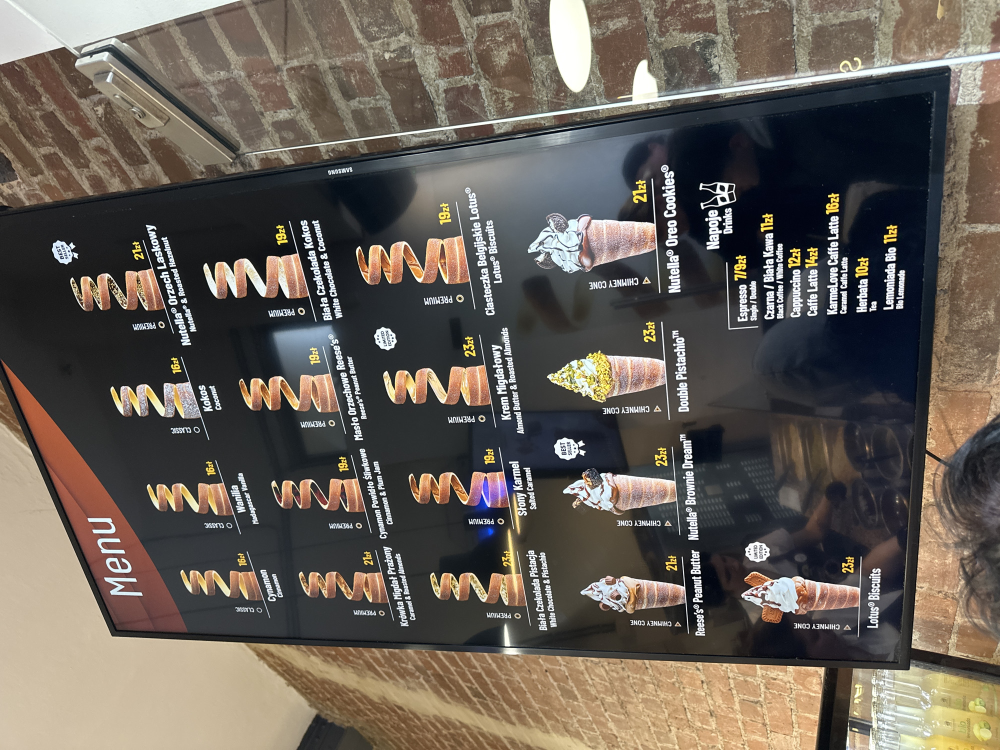 This image shows a close-up of a digital menu board displaying various dessert options and drinks. The menu features a variety of chimney cakes with flavors such as Nutella and Roasted Hazelnut, Coconut, Vanilla, Cinnamon, and several "Premium" options. Additionally, the menu includes several "Chimney Cone" flavors such as Double Pistachio, Nutella Brownie Dream, and Lotus Biscuits. A list of drink options is also present, including Espresso, Czarna / Biała Kawa, Cappuccino, Caffe Latte, KarmeLove Caffe Latte, Herbata, and Lemoniada Bio. The menu board is mounted on a wall composed of red brick. The presence of Polish text and currency (zł) on the menu suggests that the image was taken in Poland. Wrocław is a city in Poland. Therefore, it is possible the image was taken at a dessert shop in Wrocław, Poland.
