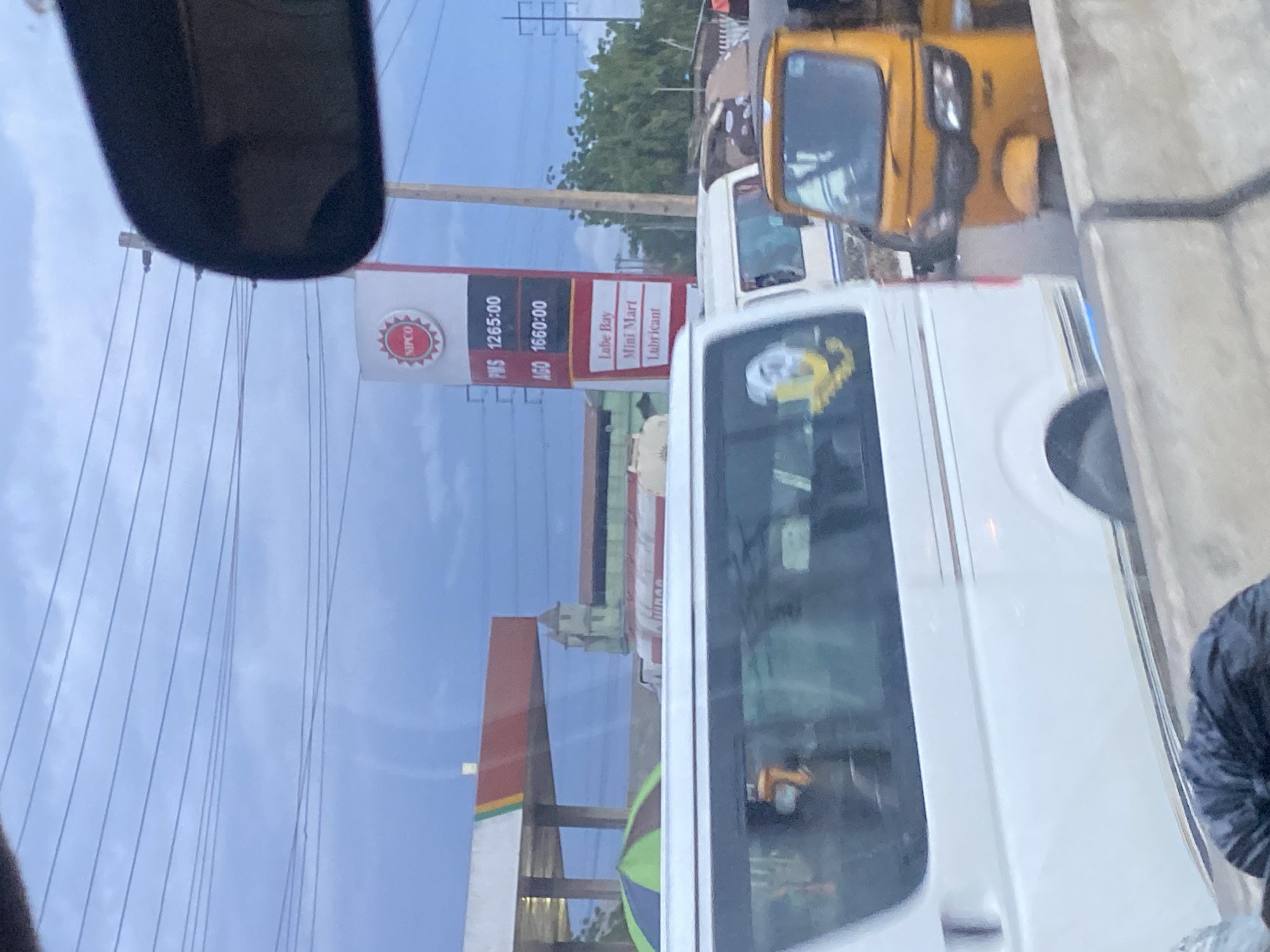 In Ikorodu, Nigeria, a NIPCO gas station sign displays fuel prices. PMS is priced at 1265.00, and AGO is 1660.00. Below these prices, the sign lists "Lube Bay," "Mini Mart," and "Lubricant." The scene is from a vehicle's perspective, showing a white van in the foreground, a yellow tricycle behind it, and other vehicles on a road. Overhead power lines are visible against a cloudy sky. In the background, a gas station canopy and a building with distinctive architectural features are present.