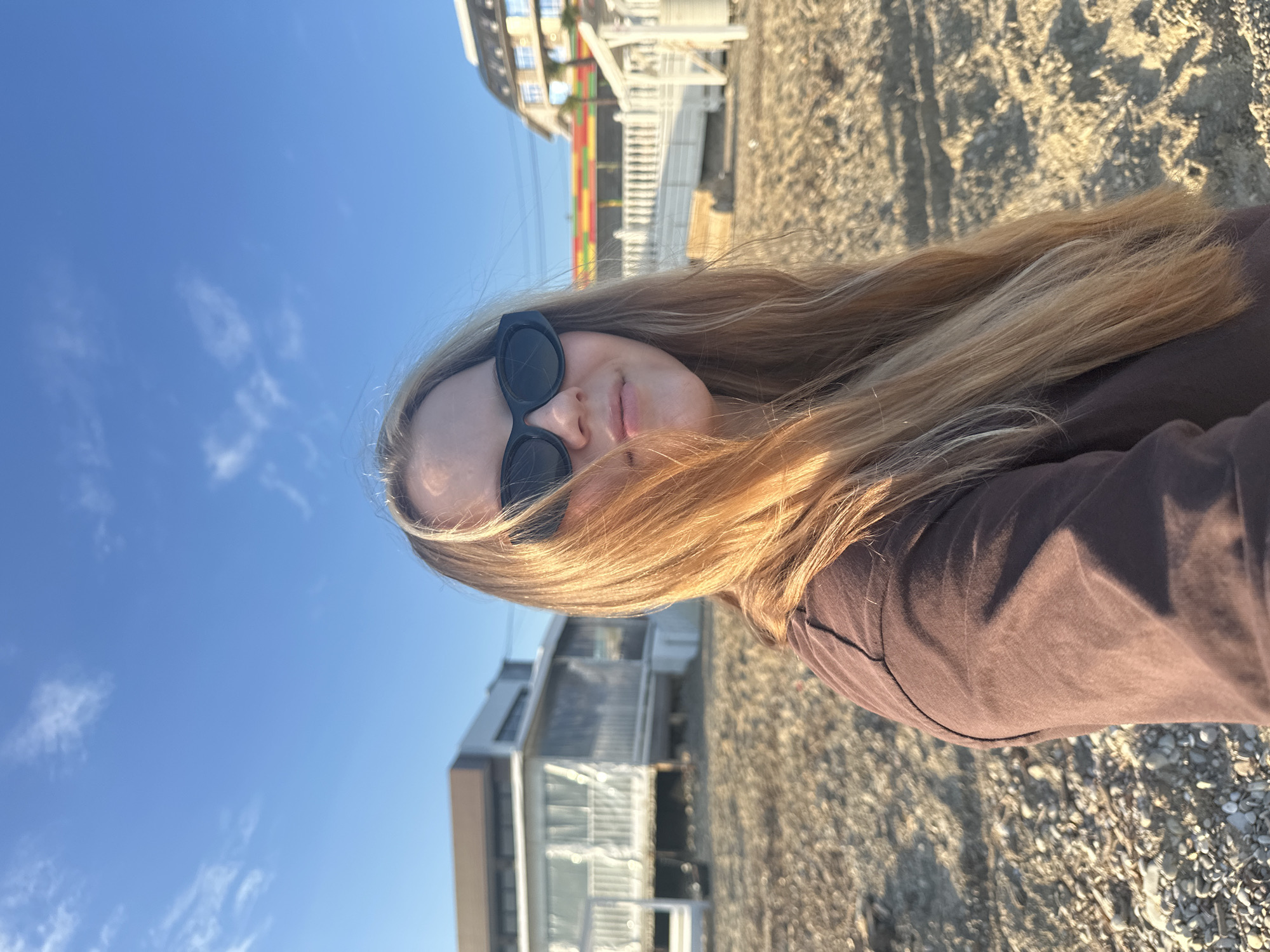 This image presents a rotated portrait of a person in an outdoor, sunny coastal environment, specifically identified as Lermontovo, Russia. The subject is a young individual, likely female, with long, flowing blonde or light brown hair. She is wearing black cat-eye sunglasses that obscure her eyes, and a dark brown or mauve long-sleeved top. Her face, partially visible from the nose down, shows a neutral to slightly relaxed expression with her lips lightly parted.
The background reveals a clear, bright blue sky with scattered wispy clouds. Several buildings are visible, suggesting a developed area, possibly resort housing or coastal residences. One building features prominent white railings and balconies, while another displays colorful horizontal stripes. The ground beneath the buildings and extending into the foreground is a mix of sand and small pebbles or rocks, consistent with a beach. The bright sunlight casts distinct, elongated shadows on the pebbly ground, indicating either morning or late afternoon. The overall scene conveys a pleasant, serene atmosphere on a sunny day by the sea. No visible text is present in the image.