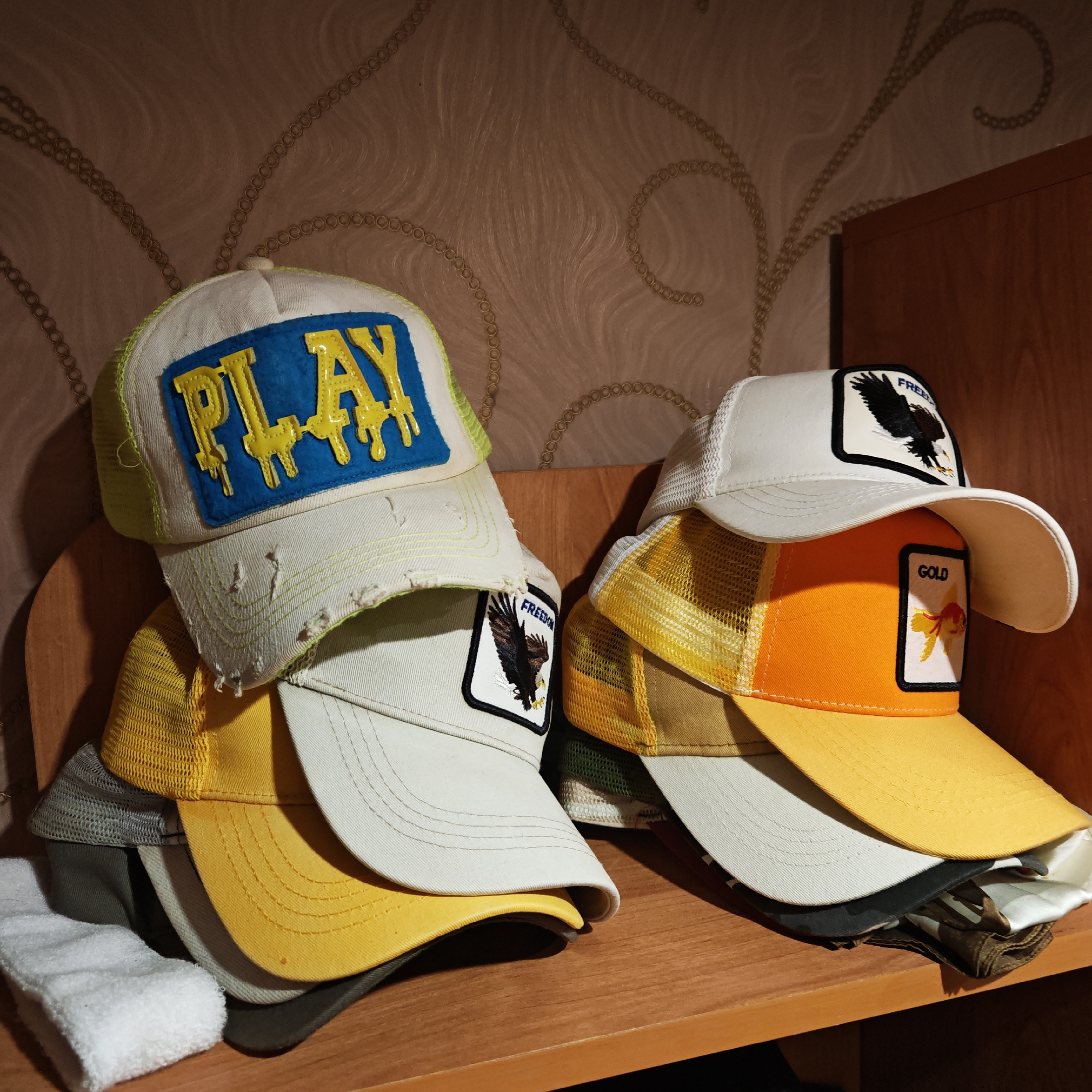 A collection of trucker hats and baseball caps is displayed on a light brown wooden shelf in Kladovitsy, Russia. The hats are arranged in two main stacks.
The stack on the left is topped by an off-white cap with a distressed, frayed brim. Its front features a prominent blue rectangular patch with the word "PLAY" in shiny, yellow, drip-effect letters. Beneath this are several other hats, including one with a white square patch depicting an eagle and the word "FREEDOM," along with various caps in shades of yellow, cream, and gray, some featuring mesh backs. A white, rolled-up towel or cloth is visible at the bottom left of this stack.
The right stack is led by a white cap with a patch of an eagle and the word "FREEDOM." Below it is an orange mesh-backed hat with a white patch featuring a goldfish and the word "GOLD." Further down, more hats in yellow, cream, and other muted tones are stacked.
The background wall is covered with wallpaper that has a repeating pattern of golden, chain-like circles forming swirling, organic designs against a light brown, subtly textured surface. The lighting appears to be artificial, casting some shadows. No people or activities are visible.