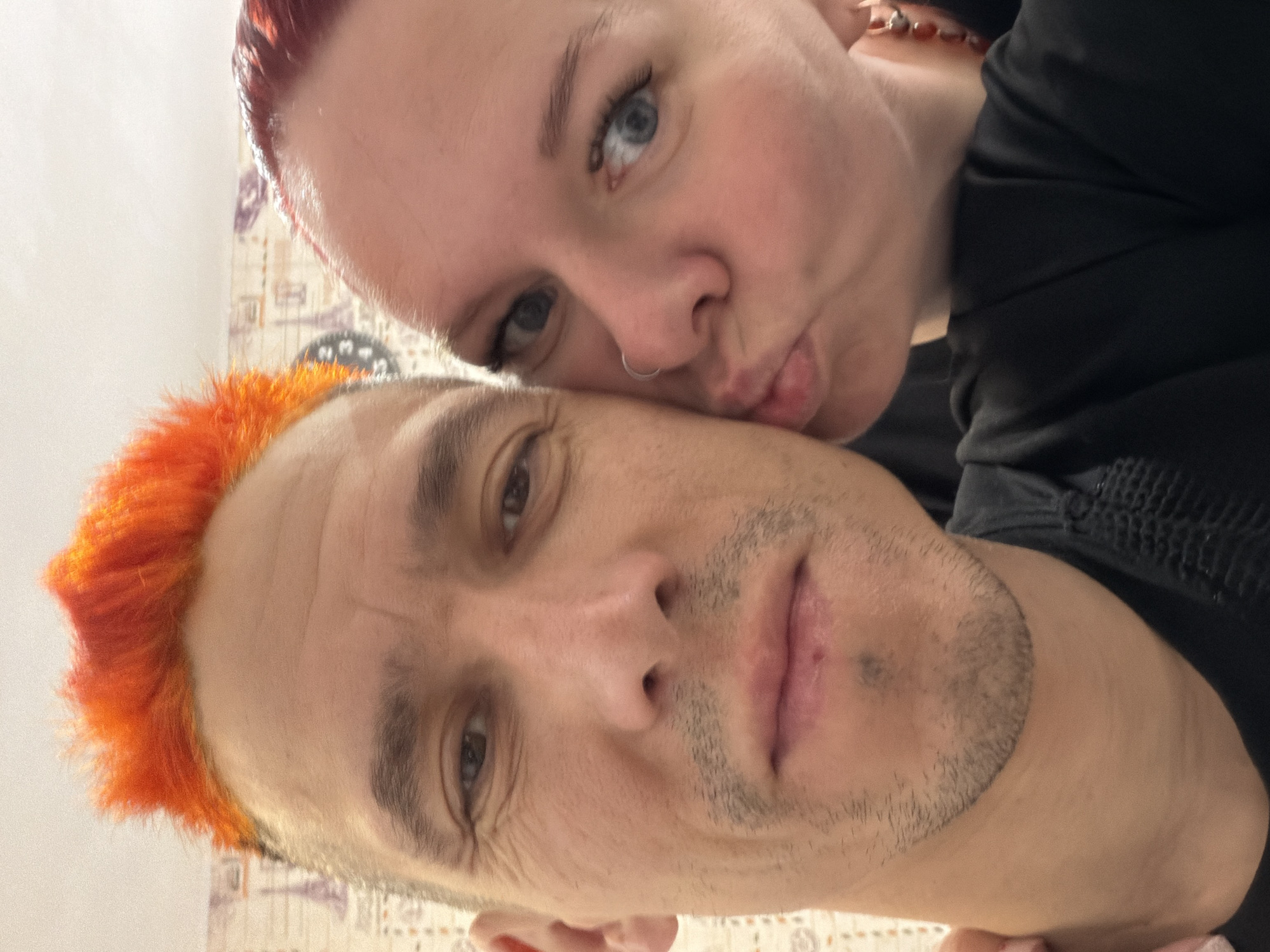 Two individuals are closely positioned in what appears to be a selfie. The person in the foreground has close-cropped orange hair. The person behind the first, slightly above and to the right, has fair skin and pursed lips. Both are wearing dark clothing. The background is indistinct, possibly showing a patterned wall. The image provides no information to determine a location in Chaikovsky, Russia.