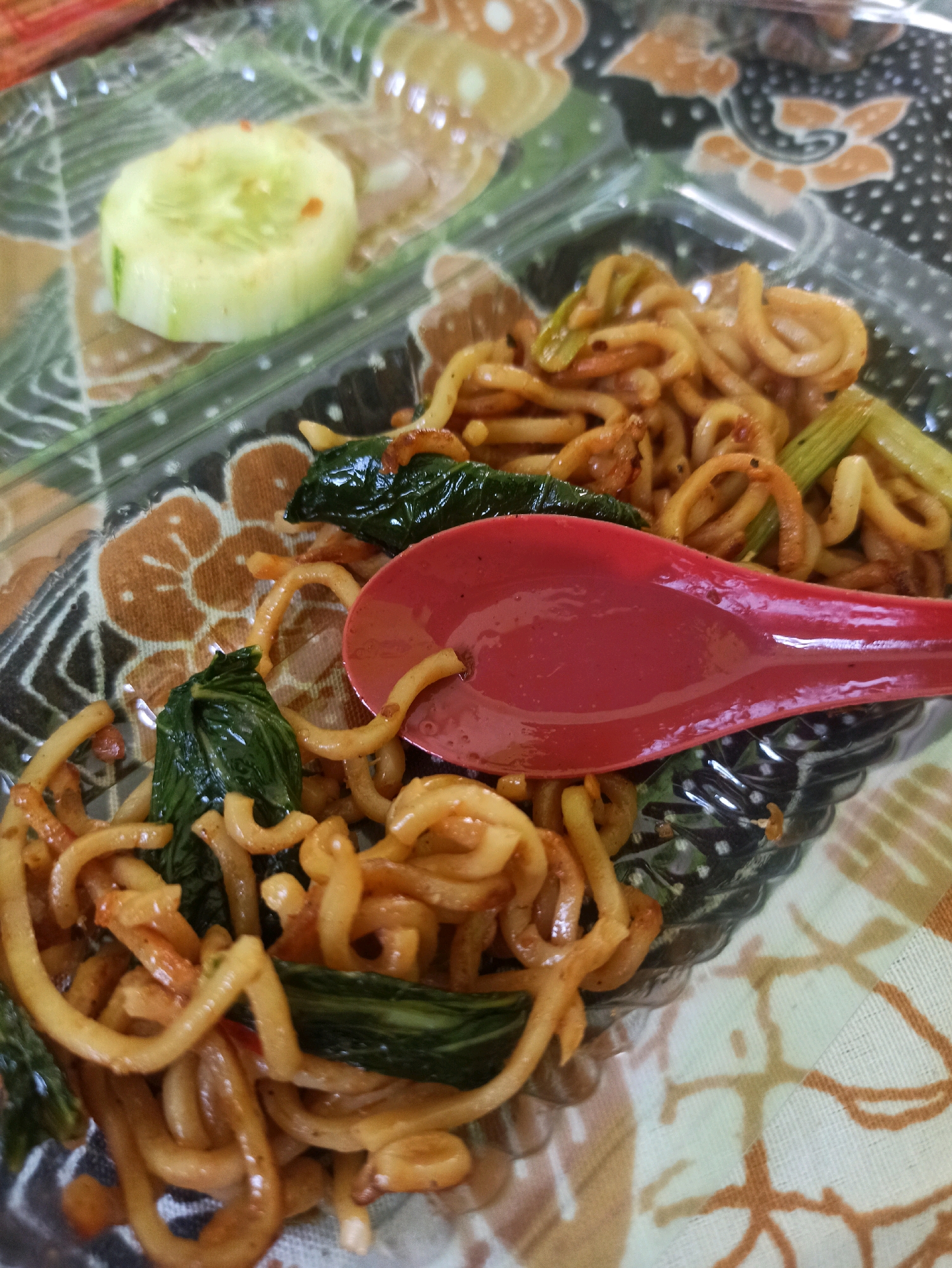 A close-up shot captures a plastic container filled with fried noodles and a slice of cucumber. The noodles are golden brown and appear to be tossed with a sauce, mixed with green leafy vegetables and possibly some thinly sliced green onions. A bright red spoon rests on top of the noodles, partially obscuring them. The container is placed on a patterned cloth, which has a light beige background with abstract brown and white floral motifs. The image is taken from a top-down perspective, focusing on the food and the container. The setting is likely a casual meal or takeout.