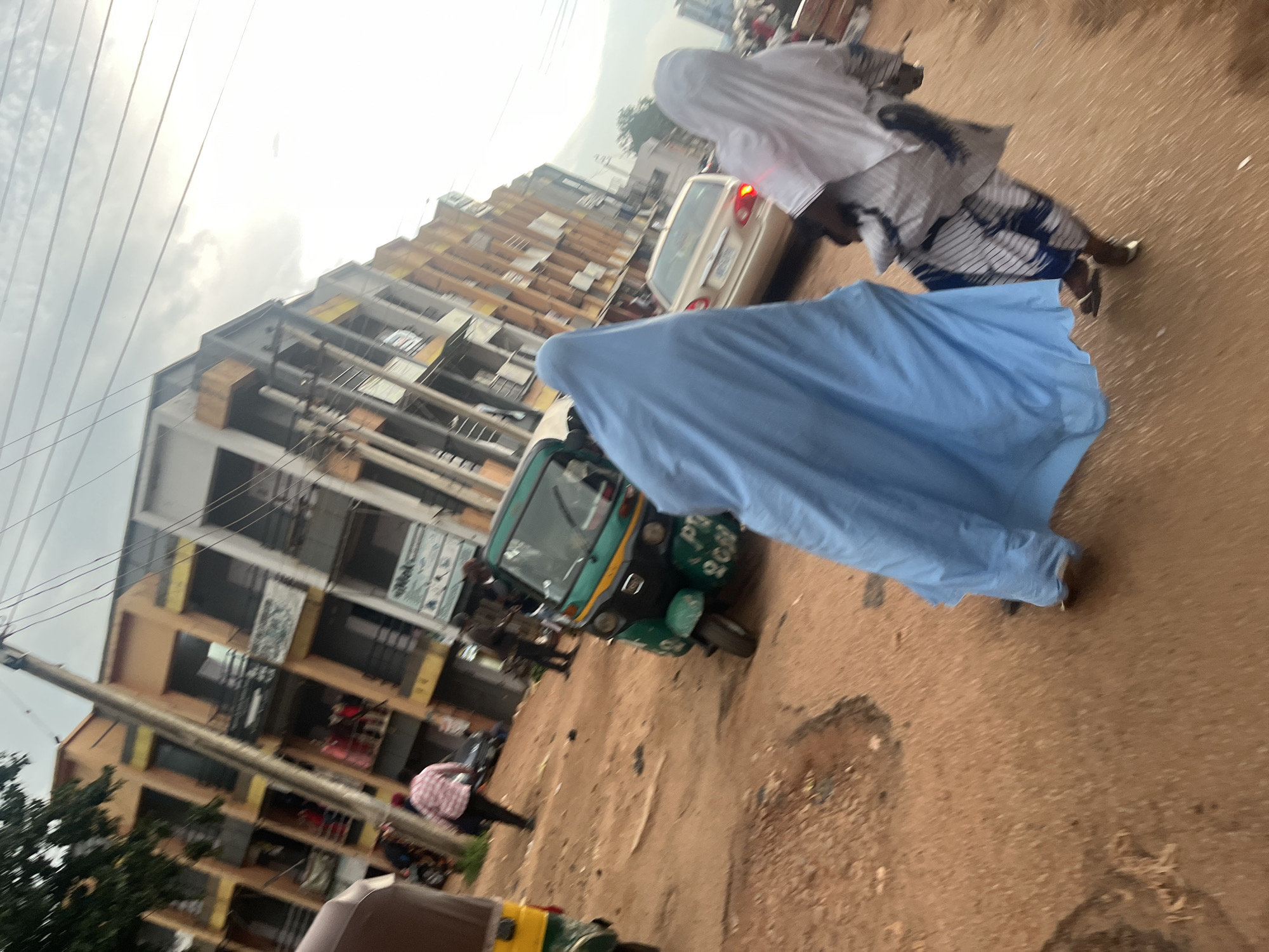 The image depicts a street scene in Jos, Nigeria, characterized by a dusty, reddish-brown dirt road. In the foreground, two individuals are walking. The person on the left is draped in a light blue garment, covering most of their body. To their right and slightly behind, another person is visible, wearing a white headscarf and a patterned dress. Their feet are in sandals.
The background features a multi-story building with a facade of orange and yellow colors. The building has several balconies with railings. Power lines crisscross the sky above. Several vehicles are present, including a green auto-rickshaw (tricycle taxi) parked on the right side of the road, and a light-colored sedan further down the street. Other people and a partial view of another auto-rickshaw are also discernible in the mid-ground. The overall impression is one of a busy urban environment.