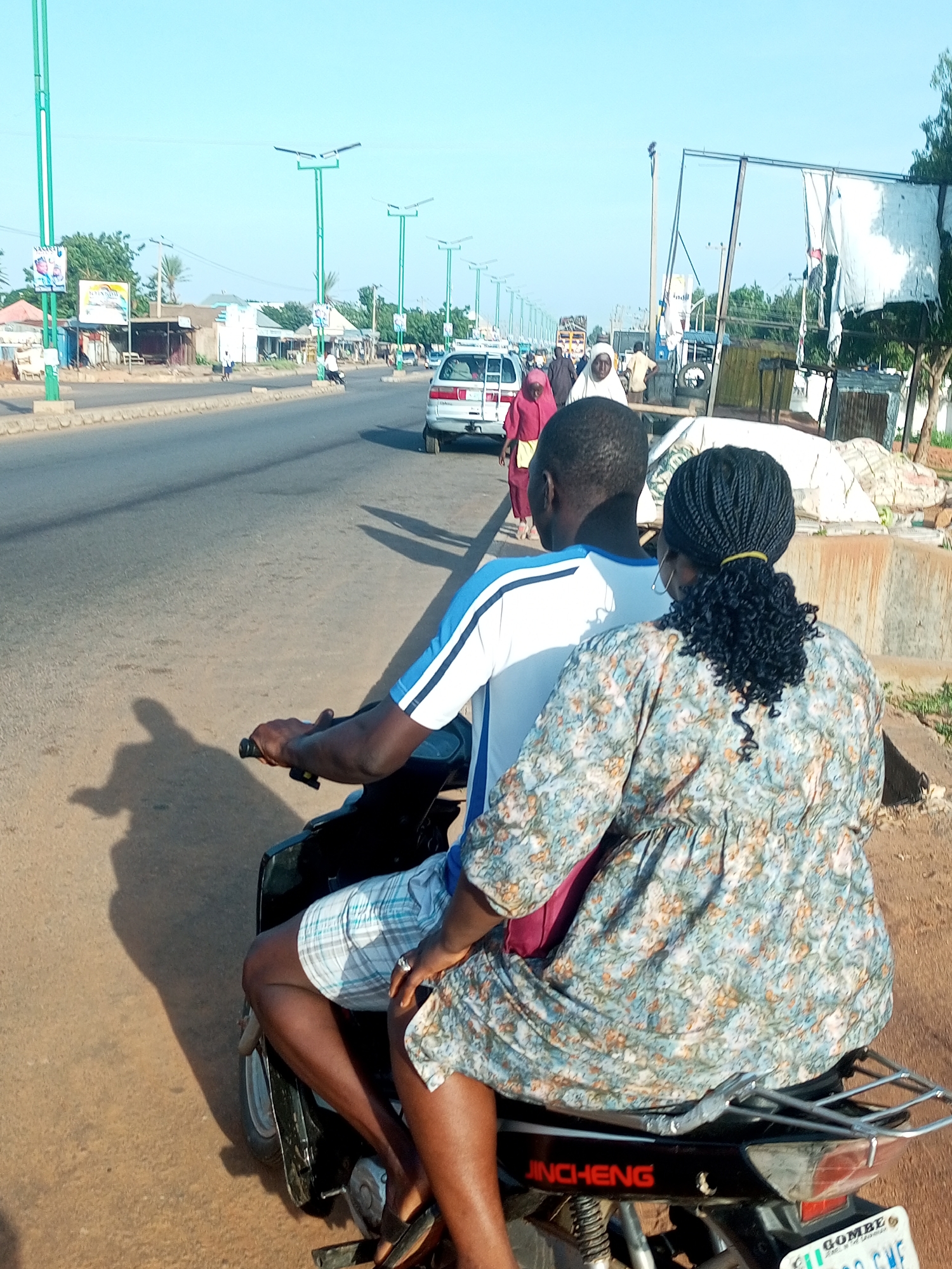 In Tempure, Nigeria, this image captures two individuals riding a motorcycle on a paved road under a clear, bright sky. The motorcycle is black and features the brand name "JINCHENG" in red lettering on its rear. The rider, a dark-skinned male, wears a blue and white striped t-shirt and plaid shorts. Seated behind him is a dark-skinned female wearing a floral-patterned dress. Her hair is braided and tied with a yellow band. The road is lined with streetlights with solar panels and buildings. In the background, various vehicles, including a white car and trucks, are visible, along with trees and scattered debris. The overall scene depicts a typical day on a busy street in Tempure.