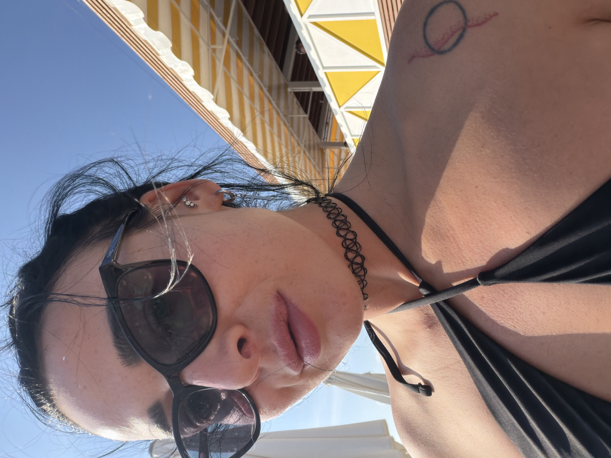 A woman wearing sunglasses and a black bikini is taking a selfie outdoors. She has dark hair pulled back and is wearing a black choker necklace. A tattoo is visible on her left shoulder. In the background, there are yellow and white awnings and a bright blue sky. The location is Shepsi, Russia, suggesting a beach or resort setting. The lighting indicates it is daytime, likely sunny.