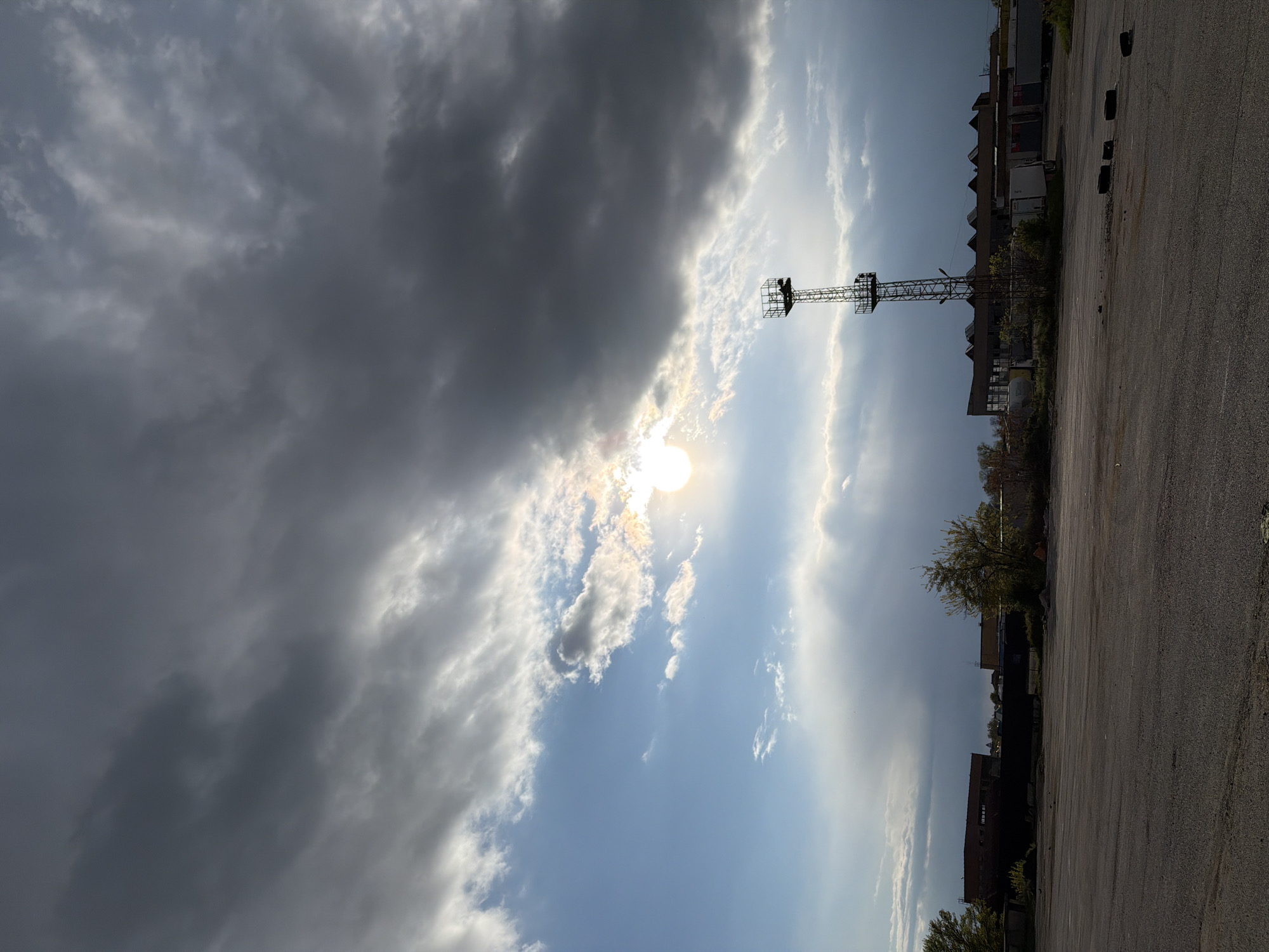 The image presents a partly cloudy sky with the sun shining through. In the foreground, there is a paved area, potentially a parking lot or open space. A metal tower-like structure stands prominently against the sky. In the background, there appear to be buildings or industrial structures. The image is oriented vertically. The city is unknown.