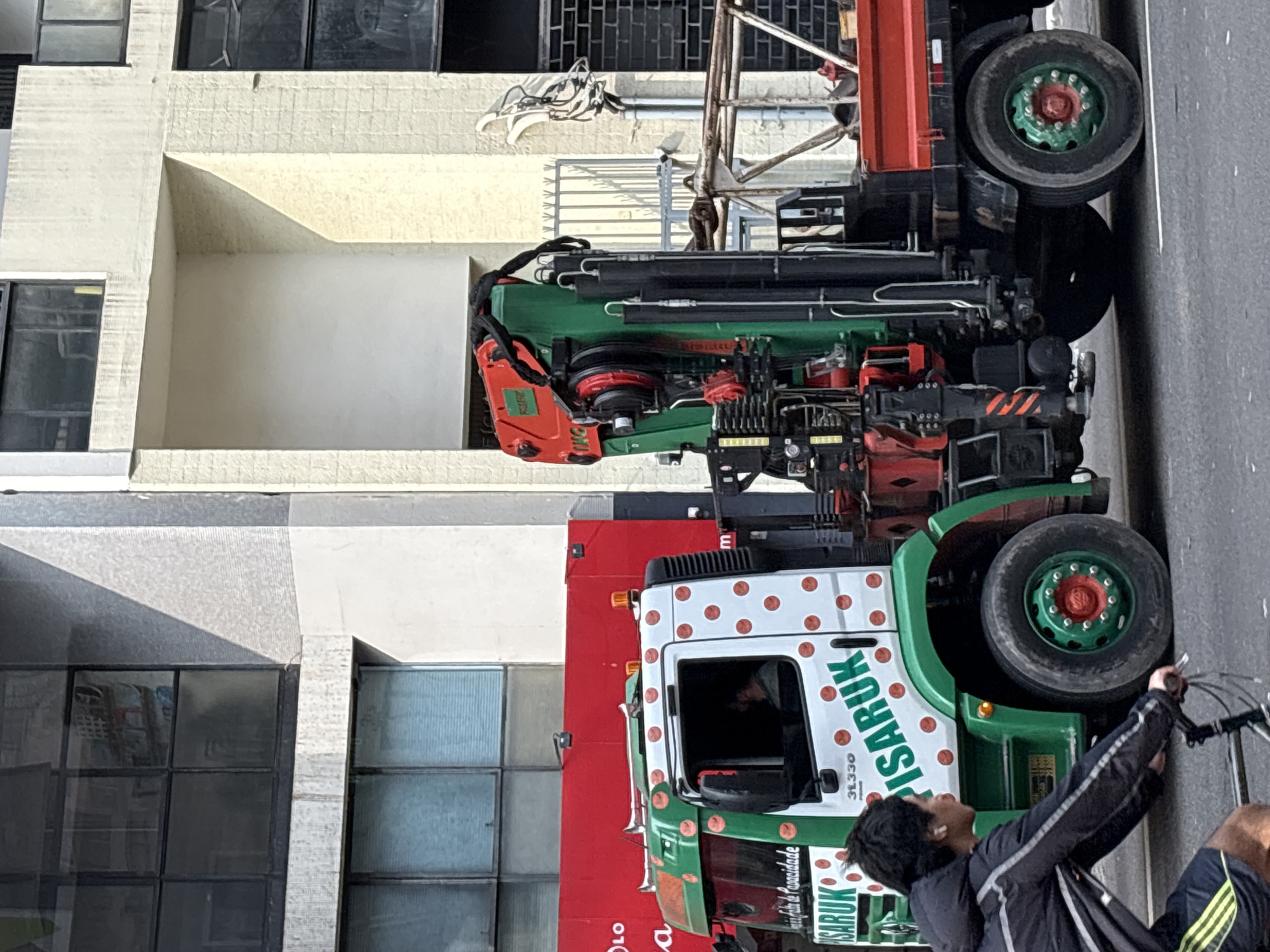 A green and white crane truck with orange polka dots is parked on a city street in São Paulo, Brazil. The truck has a large green crane attached to its back. A person is riding a bicycle in the foreground, partially obscuring the truck. The background shows a multi-story building with large windows and a red trailer. The street is paved and there are some shadows on the ground, indicating it is daytime. The building appears to be made of concrete and glass. The red trailer behind the truck has some writing on it, but it is not clearly legible.