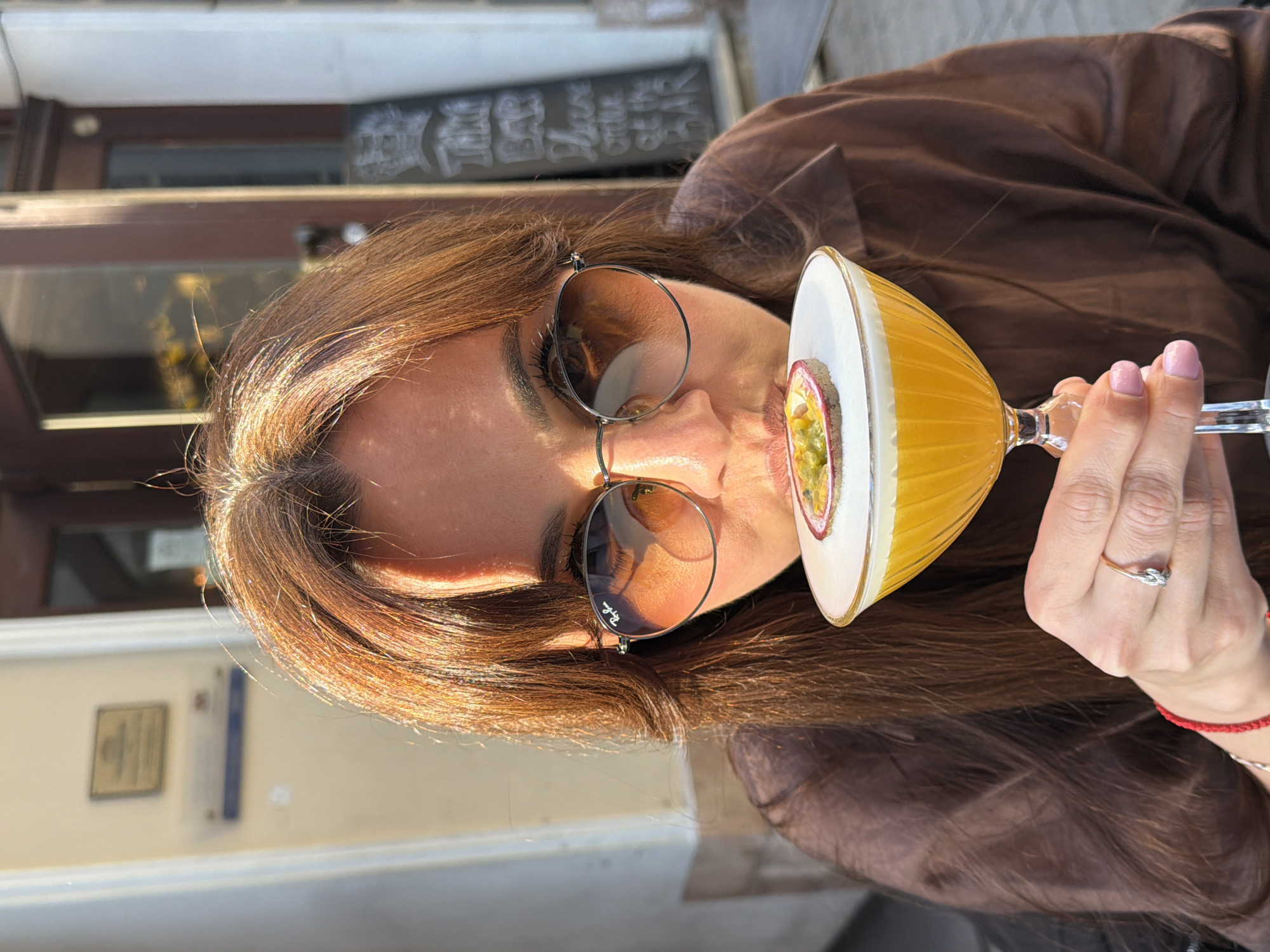 This is a close-up, side-profile image of a woman outdoors, actively drinking a cocktail on a sunny day. Her long, light brown hair is illuminated by the bright sunlight, creating warm highlights. She wears round, dark-lensed sunglasses, with the "Ray-Ban" logo clearly visible on the right lens. Her lips are pressed against the rim of a distinctive cocktail glass.
The cocktail itself is yellow or orange, presented in a ribbed glass, and features a layer of white foam on top, garnished with half a passion fruit. Her left hand, adorned with light pink nail polish, holds the stem of the glass. On her left ring finger, she wears a two-toned ring with a knot design, and a red string bracelet is visible on her wrist. She is dressed in a brown top or jacket.
The background, though somewhat blurred, indicates an outdoor cafe or bar setting. A dark wooden structure, possibly a window frame or counter, is visible behind her head. Above this, a chalkboard sign is partially visible, clearly displaying the word "BAR" and fragments of other text, appearing to say "Please order at the BAR". Further back, a lighter building wall with some indistinct plaques or signs can be seen. The scene conveys a relaxed, enjoyable atmosphere under clear, bright skies, in what the context suggests is Riga, Latvia.