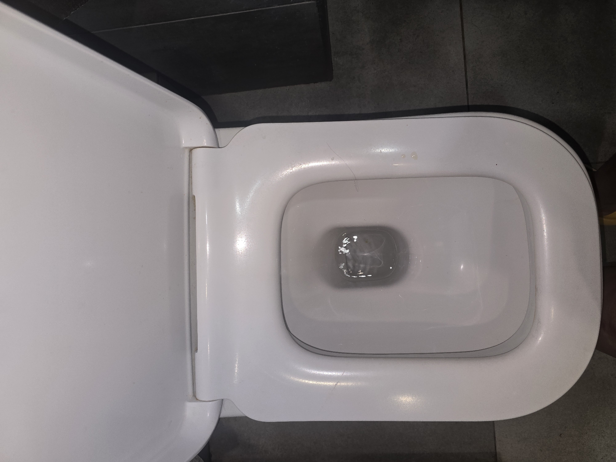 This is a top-down, close-up, eye-level shot of the interior of a white ceramic toilet bowl. The toilet seat is open and tilted upwards. The water in the bowl is slightly agitated. The surrounding floor is tiled with dark gray square tiles. The image was taken in Ulan-Ude, Russia.