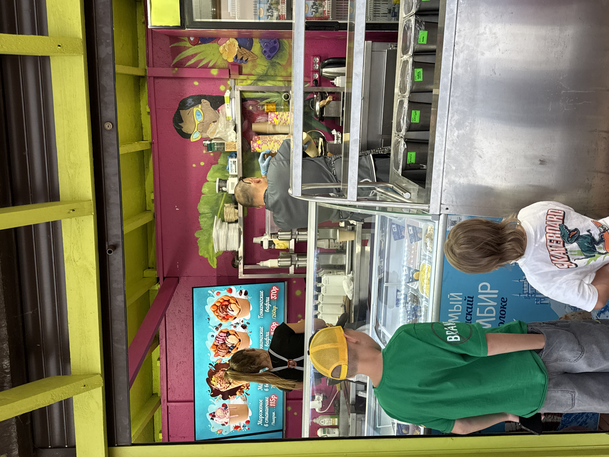 In Novofedorovka, Ukraine, an outdoor ice cream shop with a bright pink and yellow facade is depicted. The shop features a digital menu displaying various ice cream flavors and prices. Two children, one wearing a green t-shirt and the other a white t-shirt with a skateboard graphic, stand in front of a display case filled with ice cream. A female employee with her back to the camera is working behind the counter. Behind her, a male employee in a grey uniform wearing blue gloves is preparing food. The environment appears to be a sunny day, and the shop is bustling with activity.