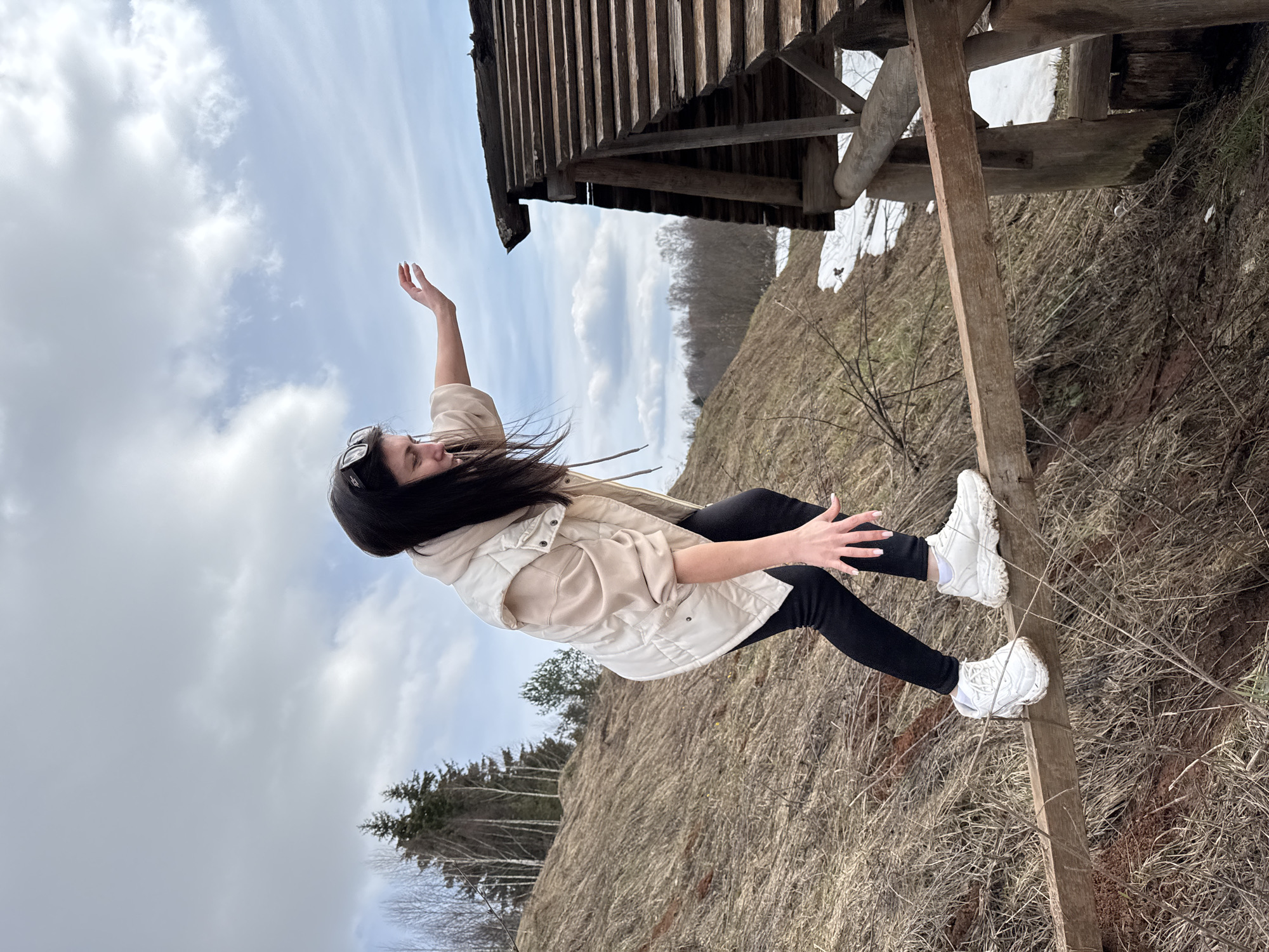 A young woman stands on a wooden structure in a rural environment in Вассята, Russia. She is wearing black pants, white sneakers, and a light beige jacket. Her dark hair is windswept, and she has one arm raised towards the cloudy sky. The structure she stands on is partially attached to a wooden building with visible snow on the ground nearby. The surrounding landscape features a grassy hillside with some trees.