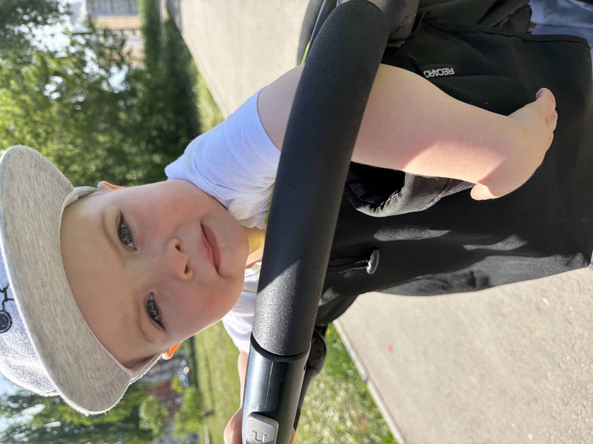 A young child, with fair skin, blue eyes, and a light gray cap, looks at the camera from inside a stroller. The child wears a white t-shirt. The stroller has a black handlebar in front of the child and a black bag attached to the stroller frame. The "RECARO" logo is visible on the bag. The background shows a paved walkway, green grass, trees, and a building. The location is Votkinsk, Russia, given the prompt's request.