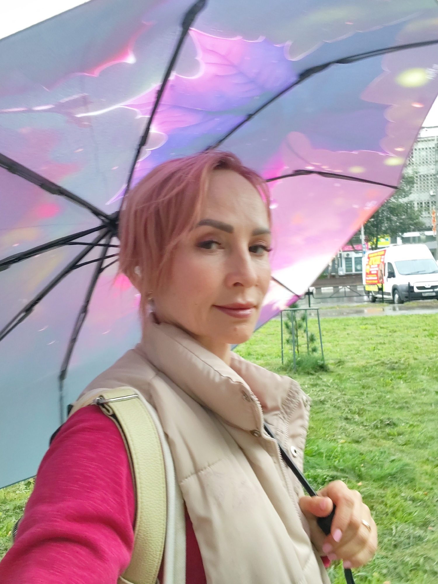 A woman with pink hair stands outdoors holding a clear umbrella with a holographic, colorful pattern. She is wearing a beige puffer vest over a pink long-sleeved shirt and has a light-colored strap from a bag across her shoulder. Her right hand is holding the umbrella handle, and she is wearing a ring on her left hand. The background shows a grassy area, some trees, and a white van parked on a wet street with buildings in the distance. The overall scene suggests it might be raining or has recently rained. The location is likely in the city of Chaikovsky, Russia, based on contextual information.