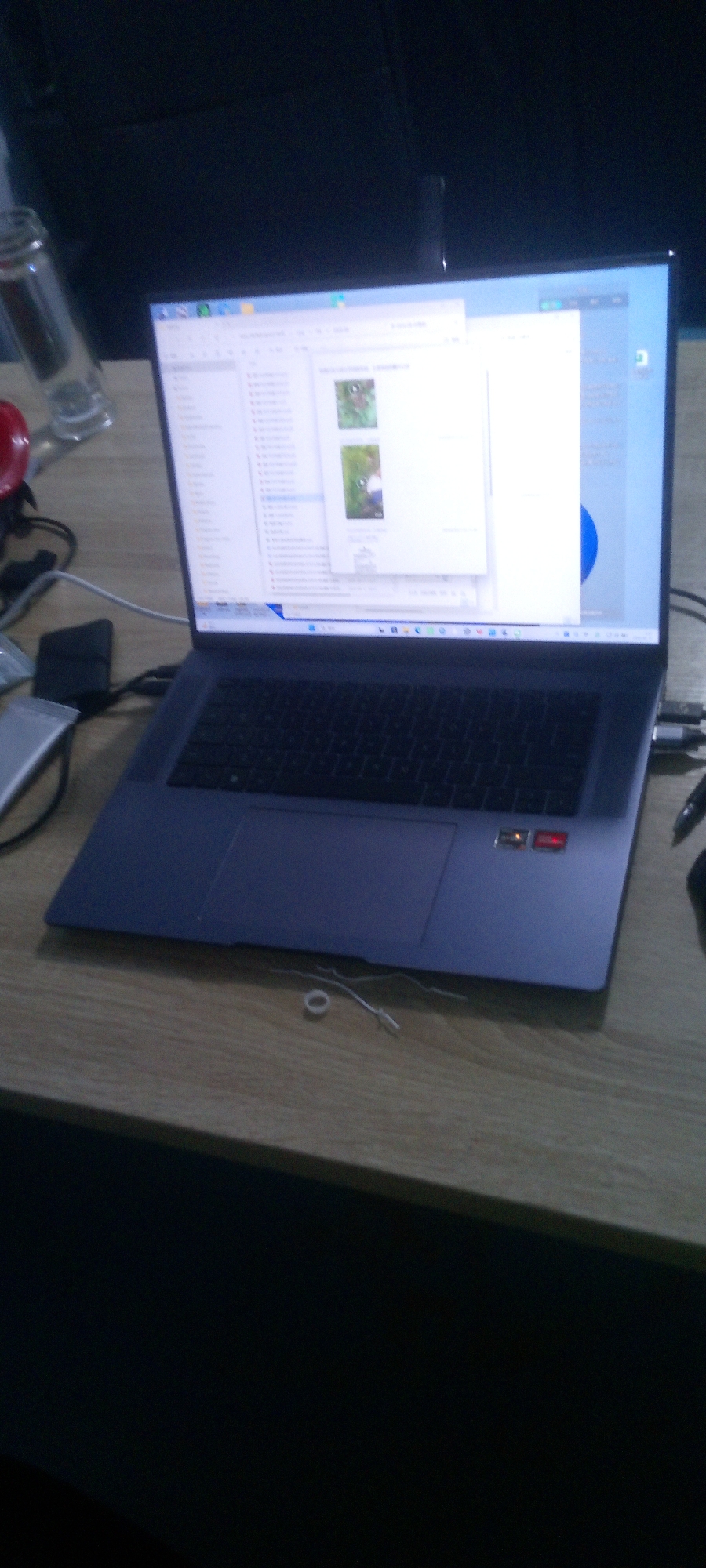 A laptop is open on a wooden desk. The screen displays a file explorer window with two small image previews. The laptop's keyboard is visible, and a mouse is partially visible on the right. To the left of the laptop, a clear glass bottle and a red object are present. Cables and a small black device are also on the desk. In the foreground, on the desk surface, a white elastic band and some thin wires are scattered. The background is dark and appears to be an indoor setting, possibly a room or office, with no discernible outdoor elements or specific location markers in Indo, Nigeria.
