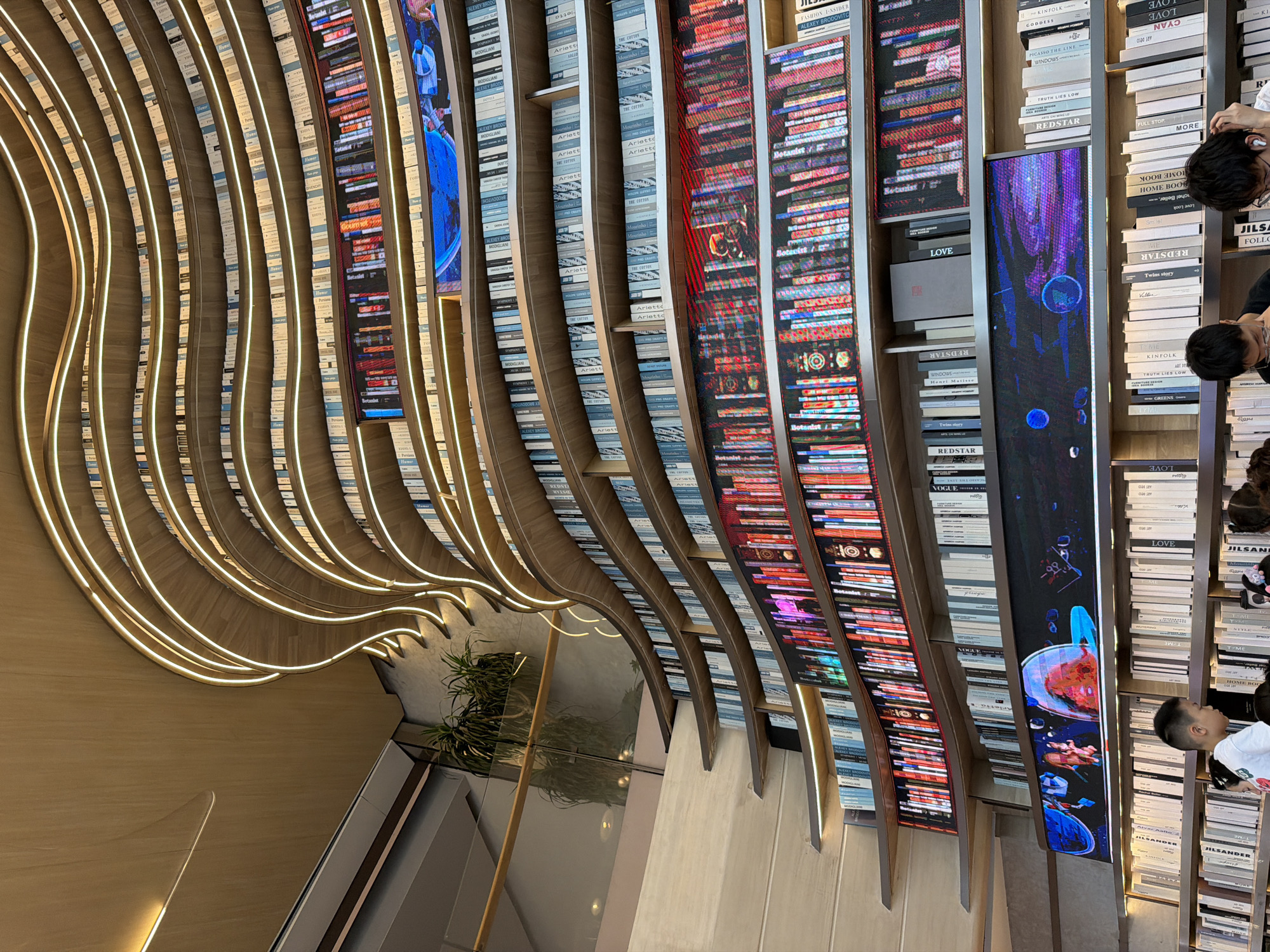 The image captures the interior of a modern library in Liangjiang, China, characterized by its striking architecture and innovative design. A towering, wave-like bookshelf dominates the frame, constructed from light-colored wood and illuminated by continuous LED strip lights that follow its undulating curves. The shelves are filled with a vast collection of books, many with light-colored spines, creating a sense of uniformity. Interspersed among the books on some shelves are digital displays showcasing vibrant, colorful imagery, including what appears to be abstract art or space scenes.
In the foreground, several individuals are present, seemingly browsing the books. A young boy in a white shirt stands prominently on the lower left, looking towards the shelves. Other individuals, including adults and possibly other children, are visible in the lower portion of the image, engaged in their activities within the library. The environment appears to be well-lit, with a warm, inviting ambiance created by the wood and lighting. The overall impression is that of a contemporary cultural space designed for reading and exploration.