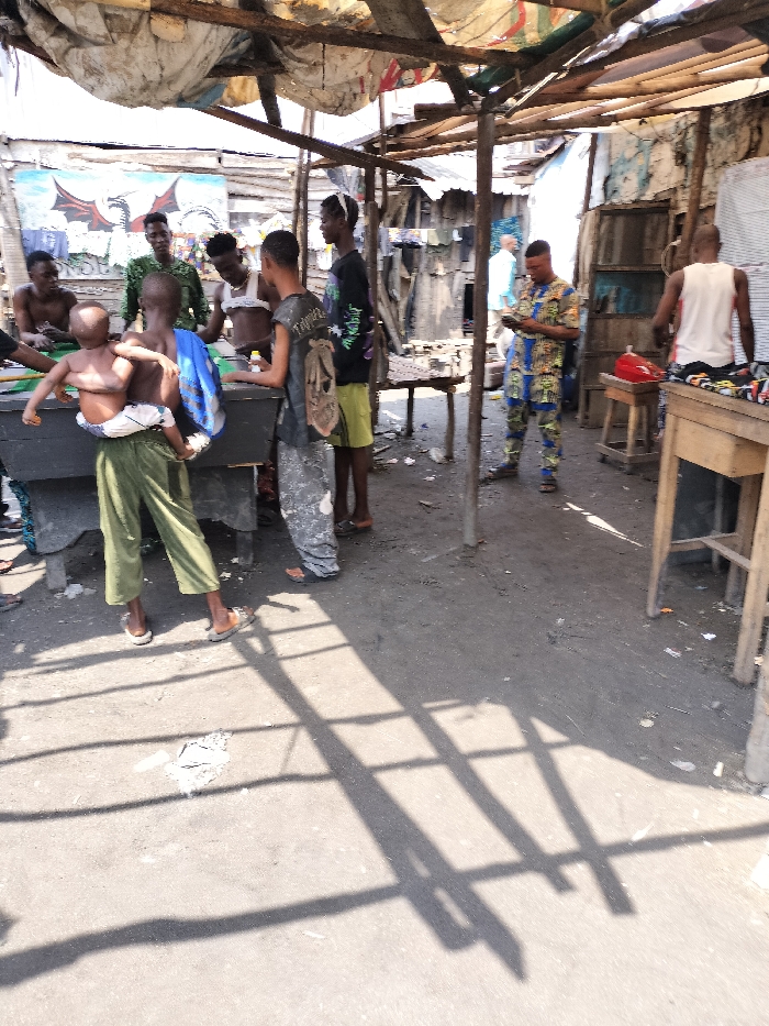 The scene depicts an outdoor gathering under a makeshift shelter in Oworonshoki, Nigeria. The shelter is constructed from rough wooden poles supporting a roof composed of corrugated metal and various patched fabric sheets. The ground is dirt or compacted earth, scattered with debris. Bright sunlight casts distinct, angular shadows from the support structure onto the ground.
Eight individuals are visible. In the foreground, a black rectangular pool table serves as a central point for a group of six males. From left to right around the table: a bare-chested male, a male adolescent in green trousers carrying an infant swaddled in white, a male in a camouflage-patterned shirt, another bare-chested male with a white fabric wrap, a male in grey camouflage trousers carrying a backpack, and a male in a black graphic t-shirt and yellow shorts. The individuals around the pool table appear to be observing or participating in a game.
In the mid-ground, a male wearing a distinctive blue, yellow, and red patterned full-body garment stands, focused on a mobile phone held in both hands. To the right, another male, seen from the back, wears a white tank top and patterned shorts while standing beside a wooden table on which a red object is visible. A light-skinned individual in a light blue shirt is partially visible further in the background, walking away. The background features several dilapidated structures constructed from wood and corrugated metal. A wall on the far left displays a vivid mural of a red and black dragon-like creature.