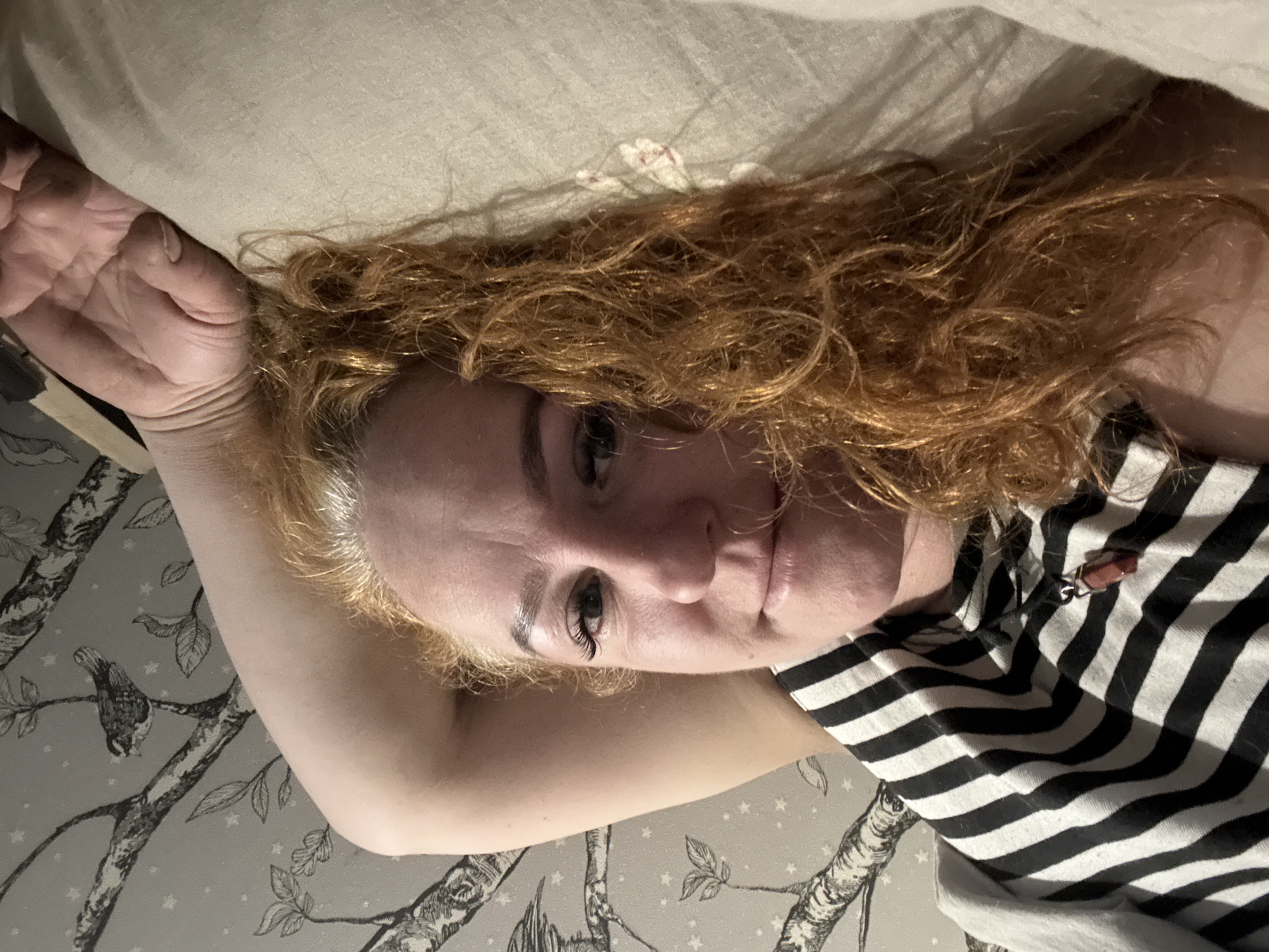 A woman with reddish curly hair lies on a bed with a beige floral patterned pillow. She has a black and white striped shirt. One arm is raised above her head. The wall is covered in wallpaper with a grey and white pattern of trees and birds. There is no indication of the location being Chaikovsky, Russia.