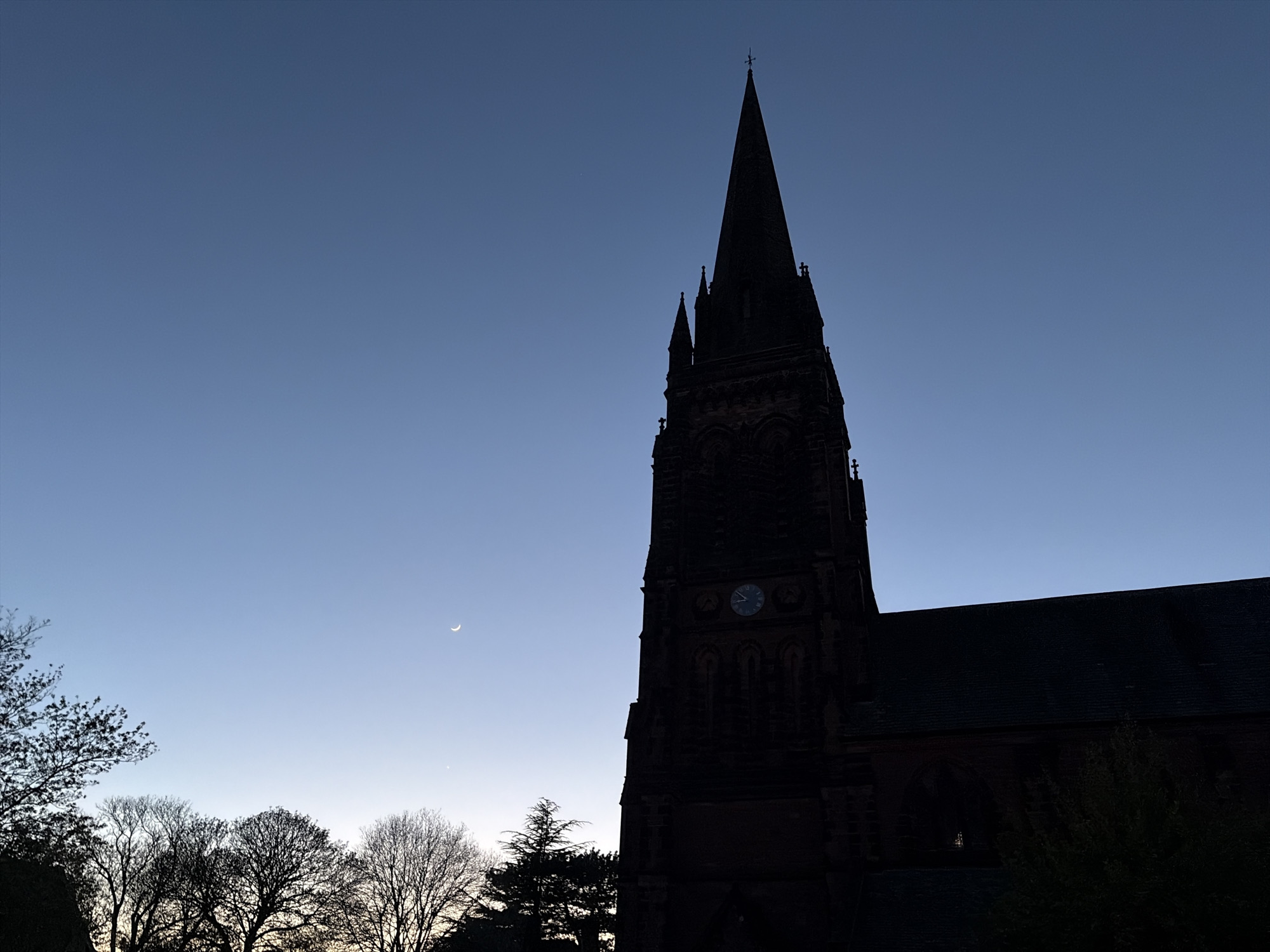 The image shows a silhouette of St. Mary's Church in Handbridge, Chester, United Kingdom. The church's spire dominates the right side of the frame. A crescent moon is visible in the blue sky to the left of the spire. Silhouetted trees line the bottom left of the image. The clock on the church tower is visible.